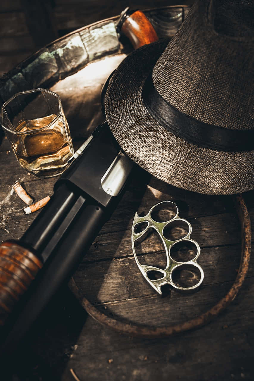 Mafia iPhone 桌布 壁紙 — 一個復古軟呢帽威士忌杯和黃銅指節套放在一張風化的木桌上，附近有一把獵槍。