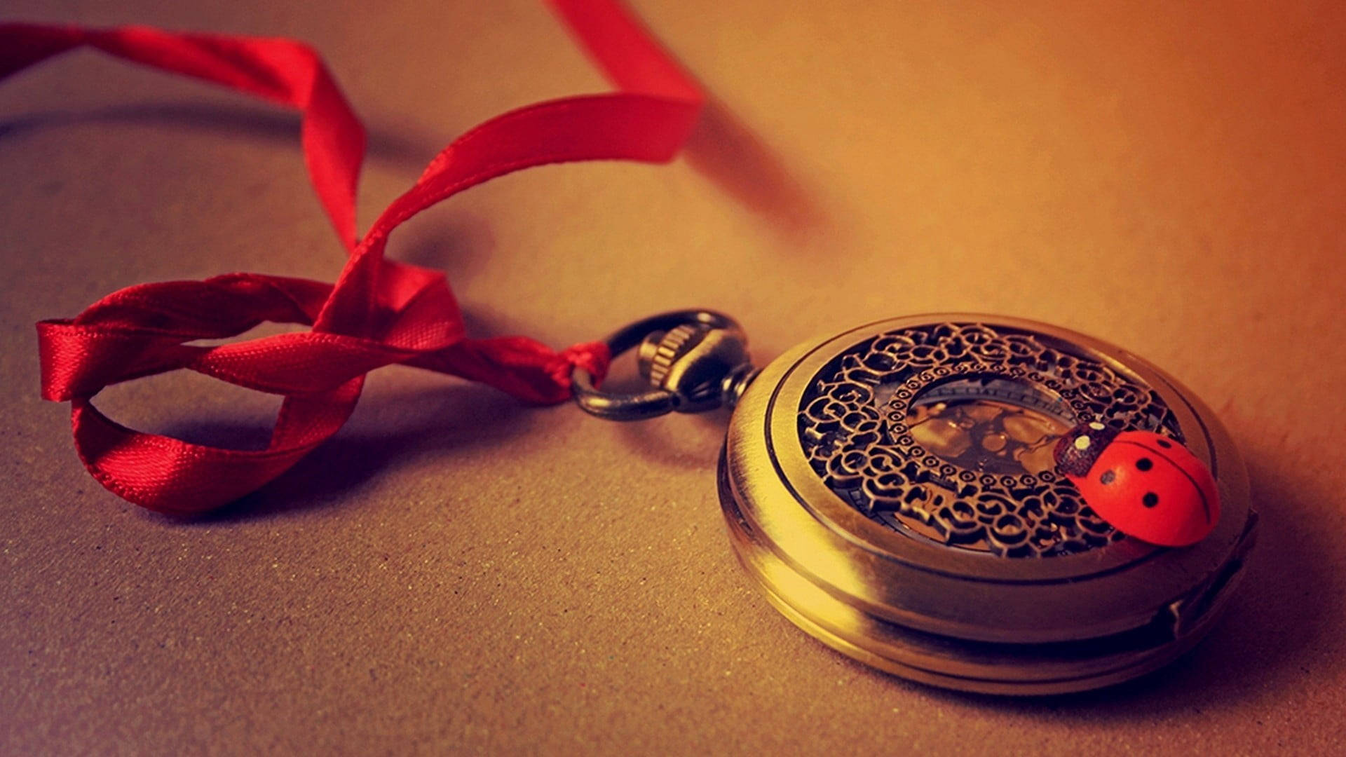 Clock 桌布 壁紙 — 一個復古黃銅懷錶，帶有精緻的鏤空花絲和紅色瓢蟲吊飾，旁邊是一條飄逸的紅色絲帶，放在溫暖的棕色表面上。