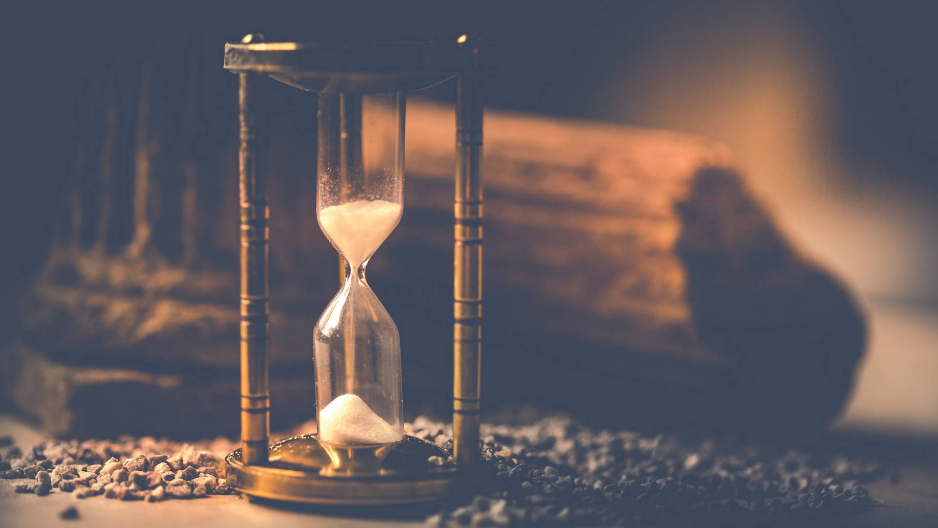Hourglass 桌布 壁紙 — 一個復古黃銅沙漏，流沙在礫石表面上，旁邊有一根模糊的木頭，在溫暖昏暗的燈光下。