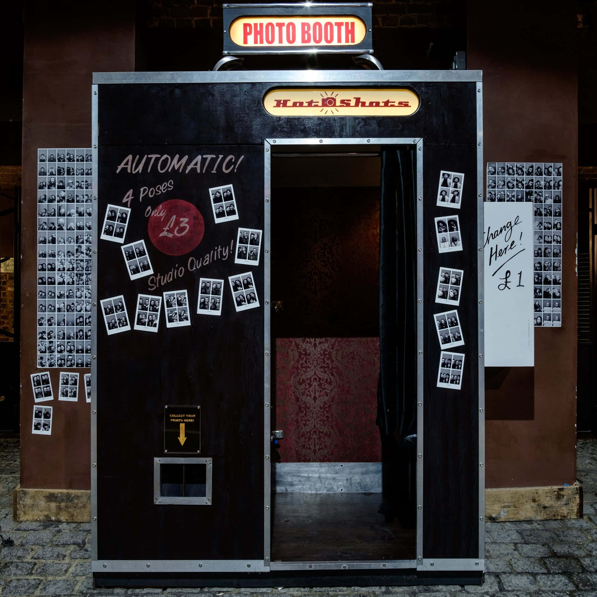 Photo Booth 桌布 壁紙 — 一個復古黑色照相亭，帶有紅色霓虹燈標誌，門上散落著即影即有照片，矗立在鵝卵石街道上。