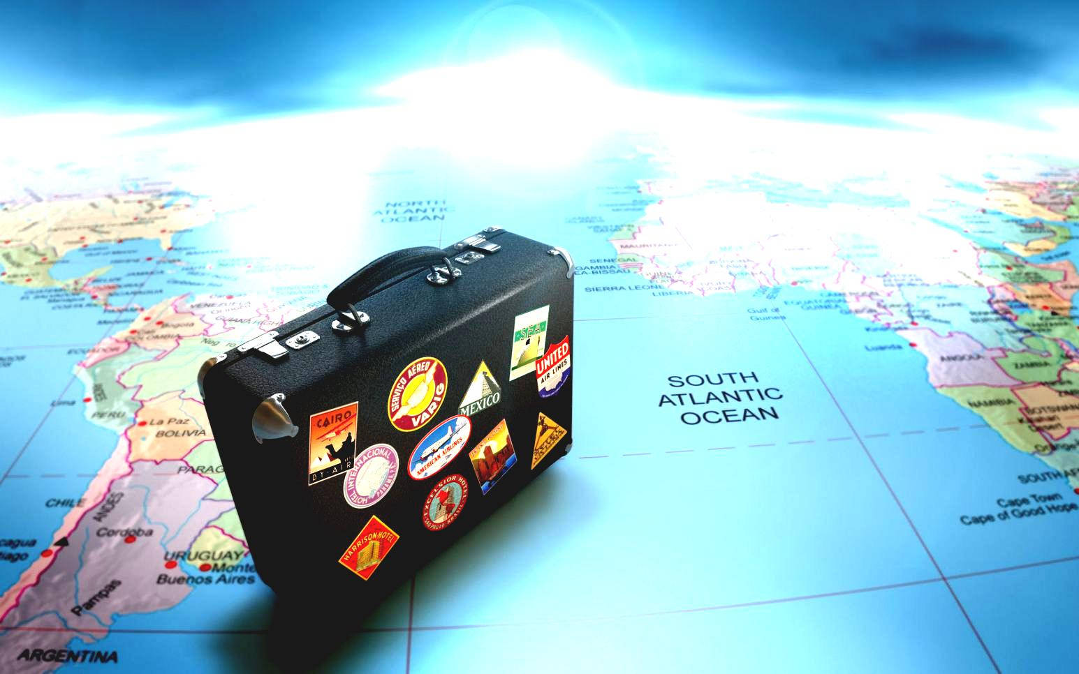Travel HD 桌布 壁紙 — 一個復古黑色行李箱，上面貼滿色彩繽紛的旅行貼紙，放在世界地圖上，上方有明亮的陽光照射。