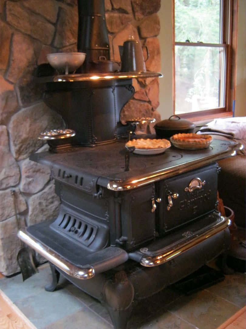 Stove 桌布 壁紙 — 一個復古黑色鑄鐵柴火爐，帶有黃銅飾邊，放置在石壁爐中，上面有餡餅，窗外顯示外面的綠色植物。