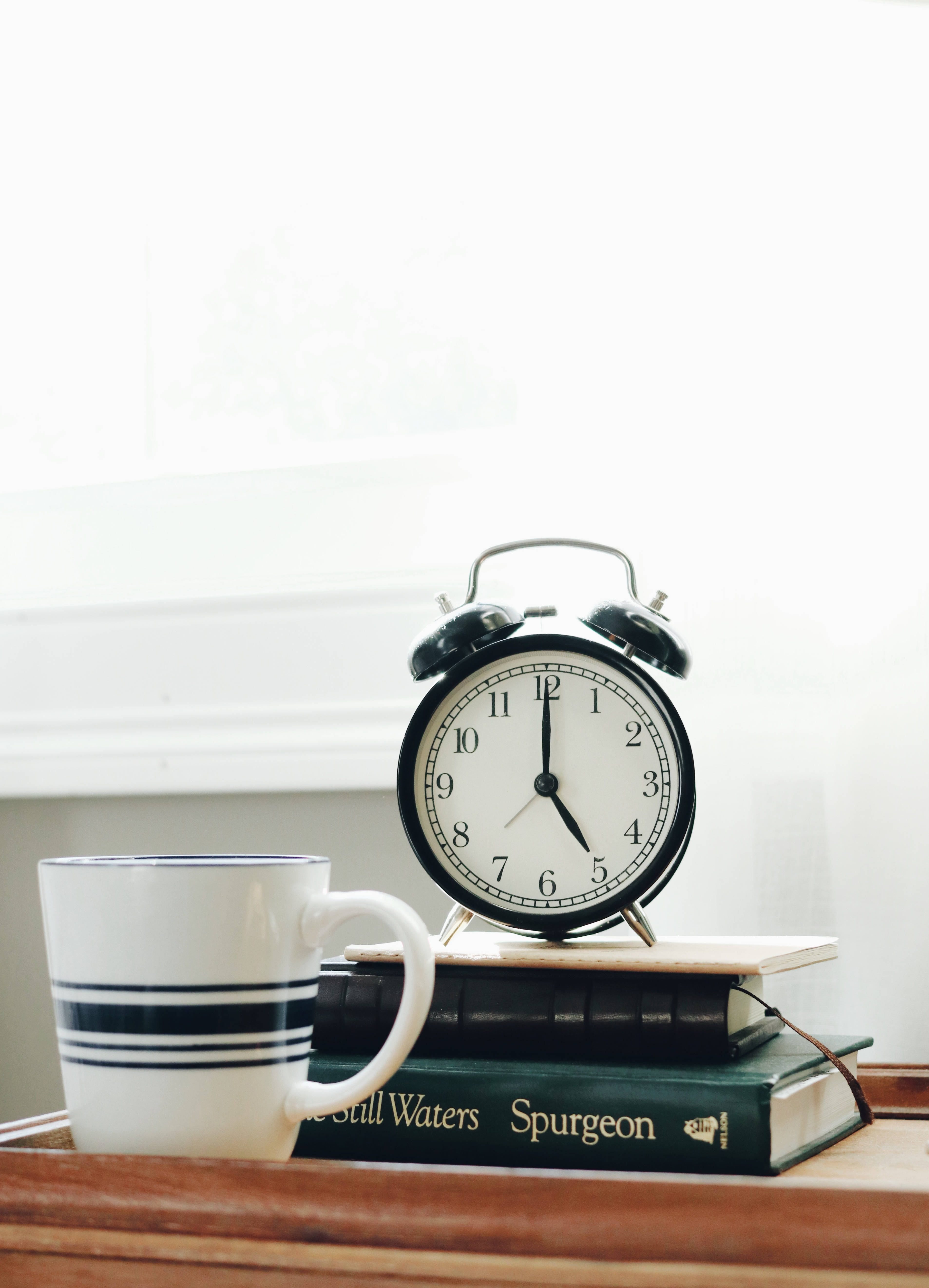 Clock 桌布 壁紙 — 一個復古黑色鬧鐘放在書本上，旁邊有一個條紋馬克杯，置於木窗台上，沐浴在柔和日光中。