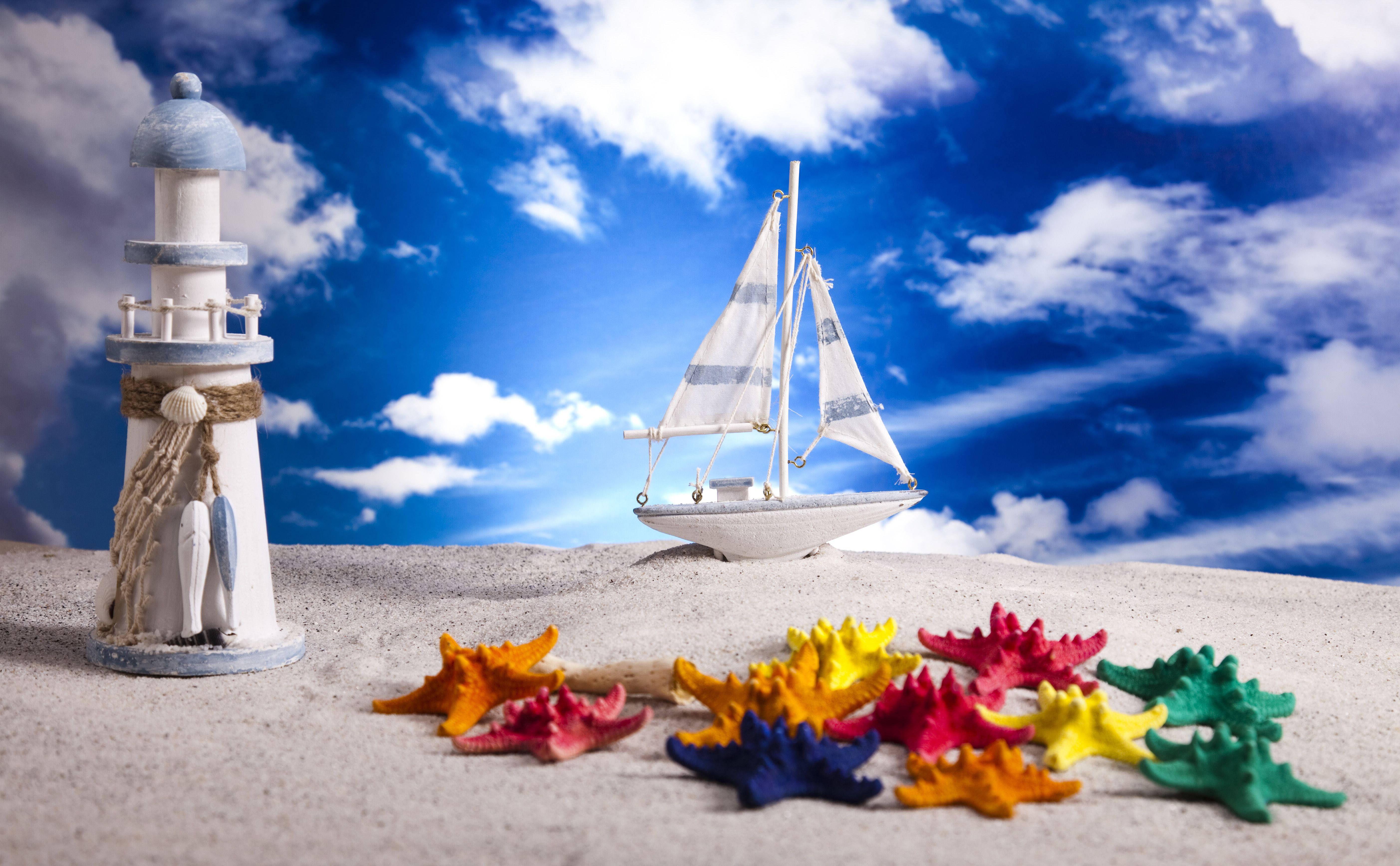 Beach Desktop 桌布 壁紙 — 一個微型燈塔和帆船坐在沙灘上，旁邊有色彩繽紛的海星，在充滿活力的藍天和 fluffy 雲朵下。