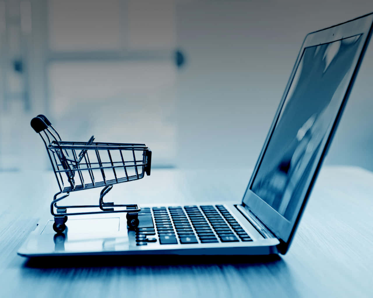 E Commerce 桌布 壁紙 — 一個微型購物車放在筆記型電腦鍵盤上，處於冷色調藍色單色辦公室環境中。