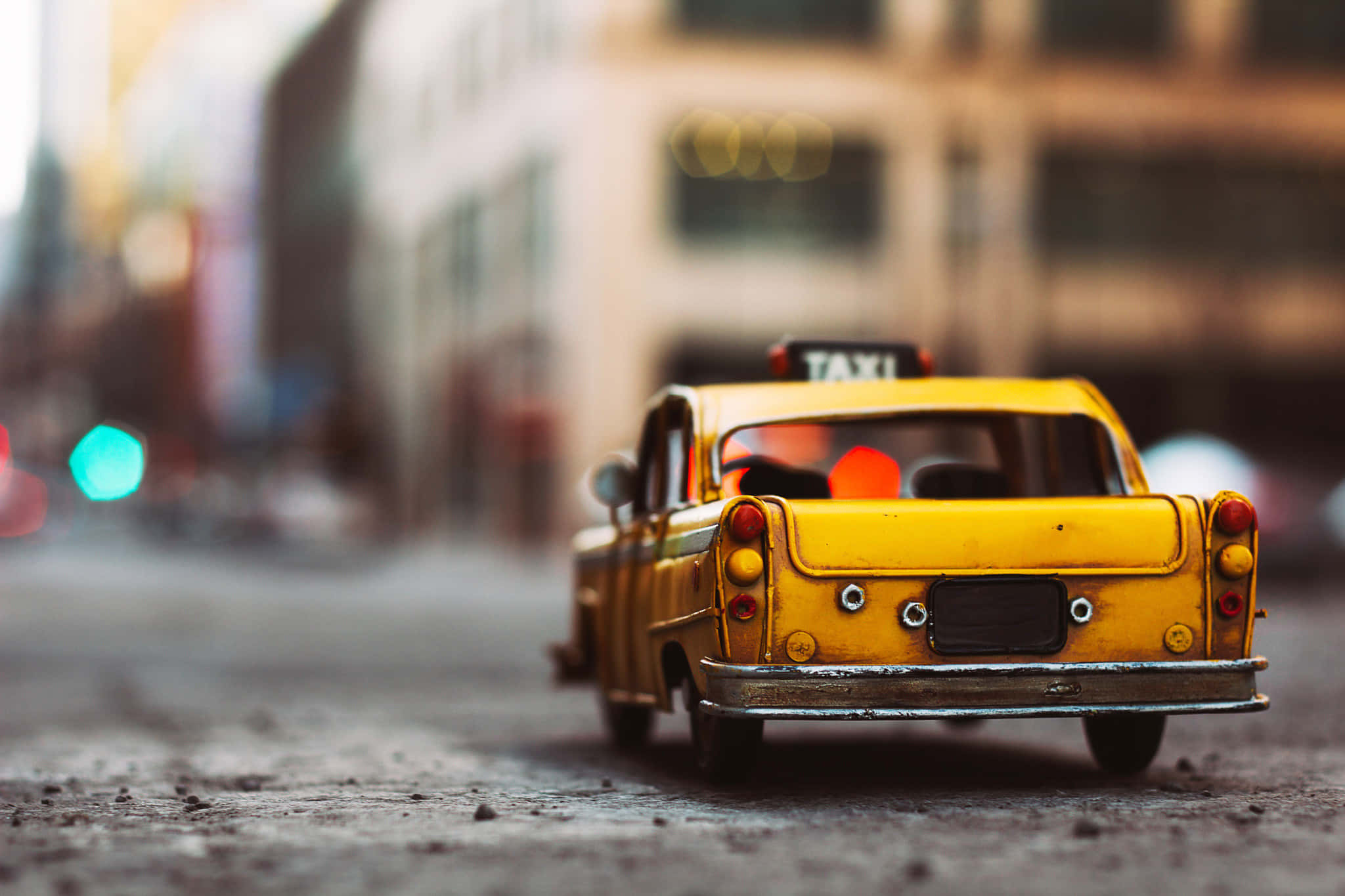 taxi 桌布 壁紙 — 一個微型黃色計程車玩具放在一條粗糙的街道上，背景是模糊的城市燈光和建築物。