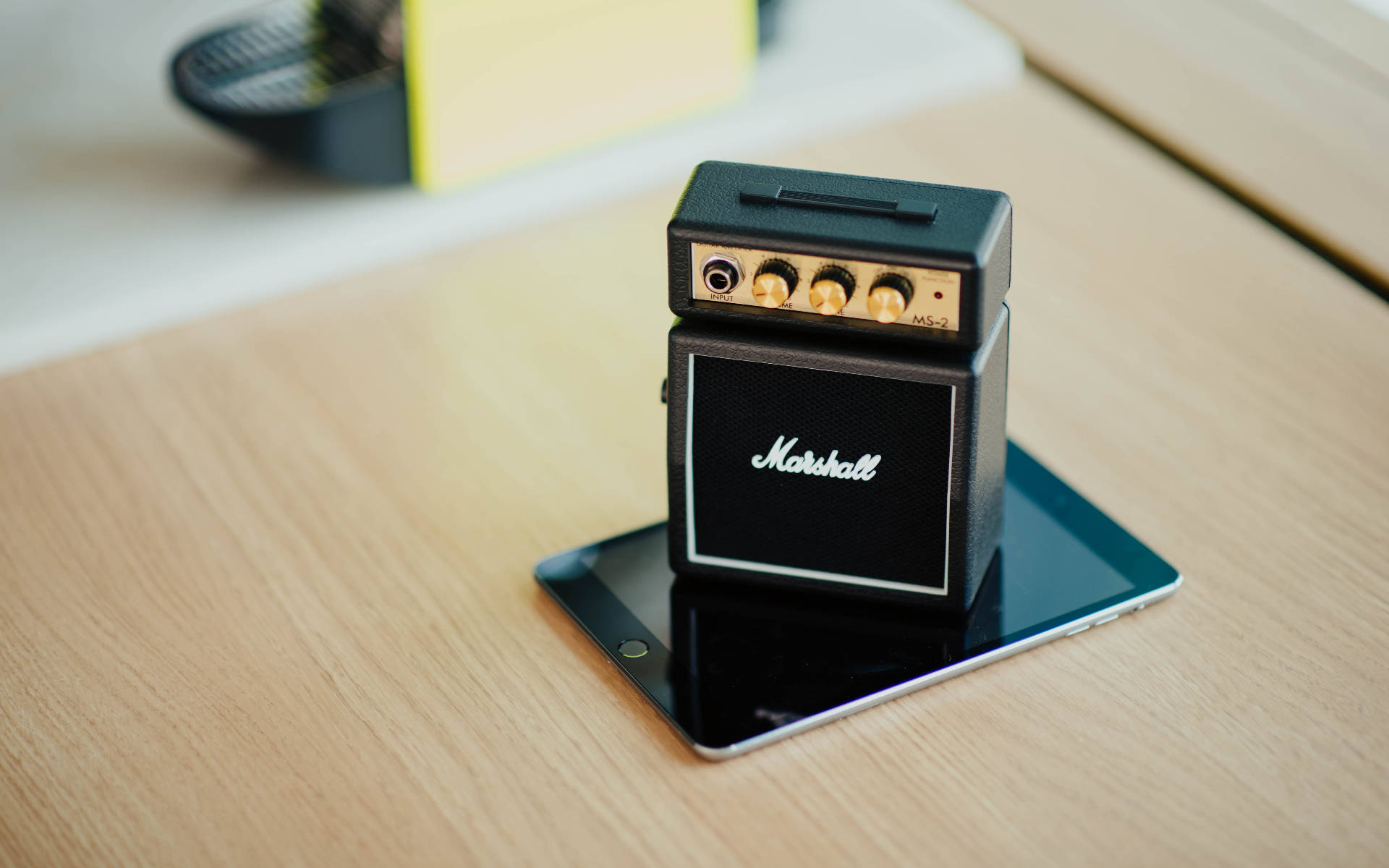 Marshall 桌布 壁紙 — 一個微型黑色Marshall吉他音箱放在一台纖薄的平板電腦上，置於淺色木桌上，柔和自然光線照射。