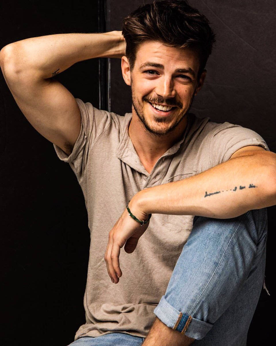 Grant Gustin 桌布 壁紙 — 一個微笑、有刺青和鬍鬚的男人，在深色背景前穿著米色襯衫和牛仔褲，隨意地擺姿勢。