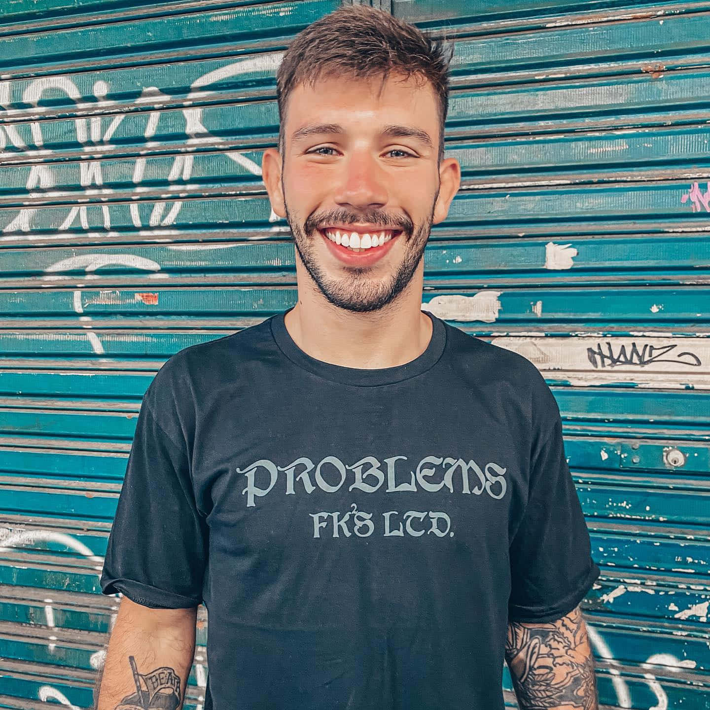 Smile 桌布 壁紙 — 一個微笑、有刺青的男人，身穿印有 'PROBLEMS FK'S LTD.' 字樣的黑色T恤，站在一個被塗鴉覆蓋的藍綠色金屬百葉門前。