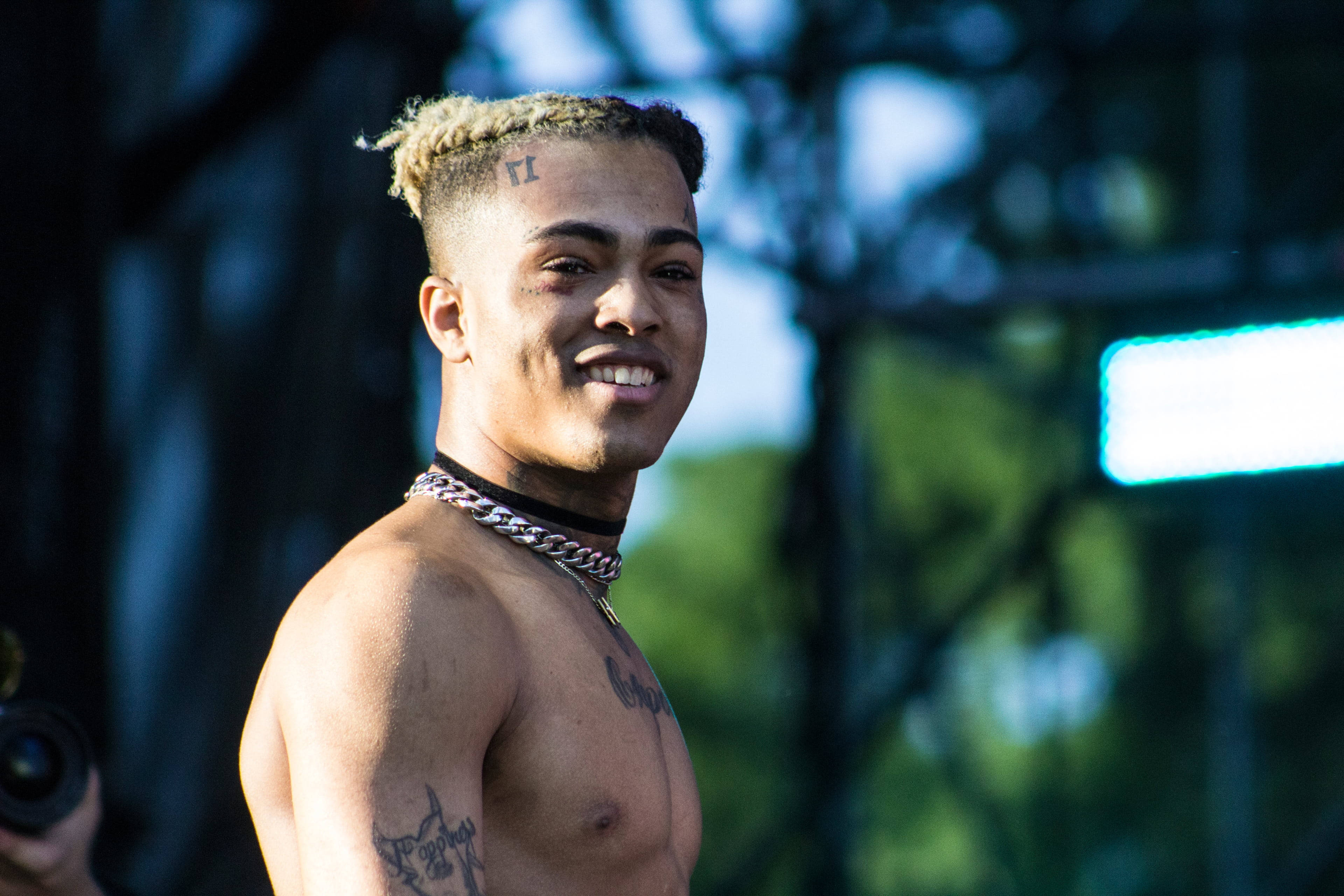 xxxtentacion 電腦 桌布 壁紙 — 一個微笑、赤膊、編織頭髮和紋身的男人，在明亮的舞台燈光下於戶外音樂會擺姿勢。