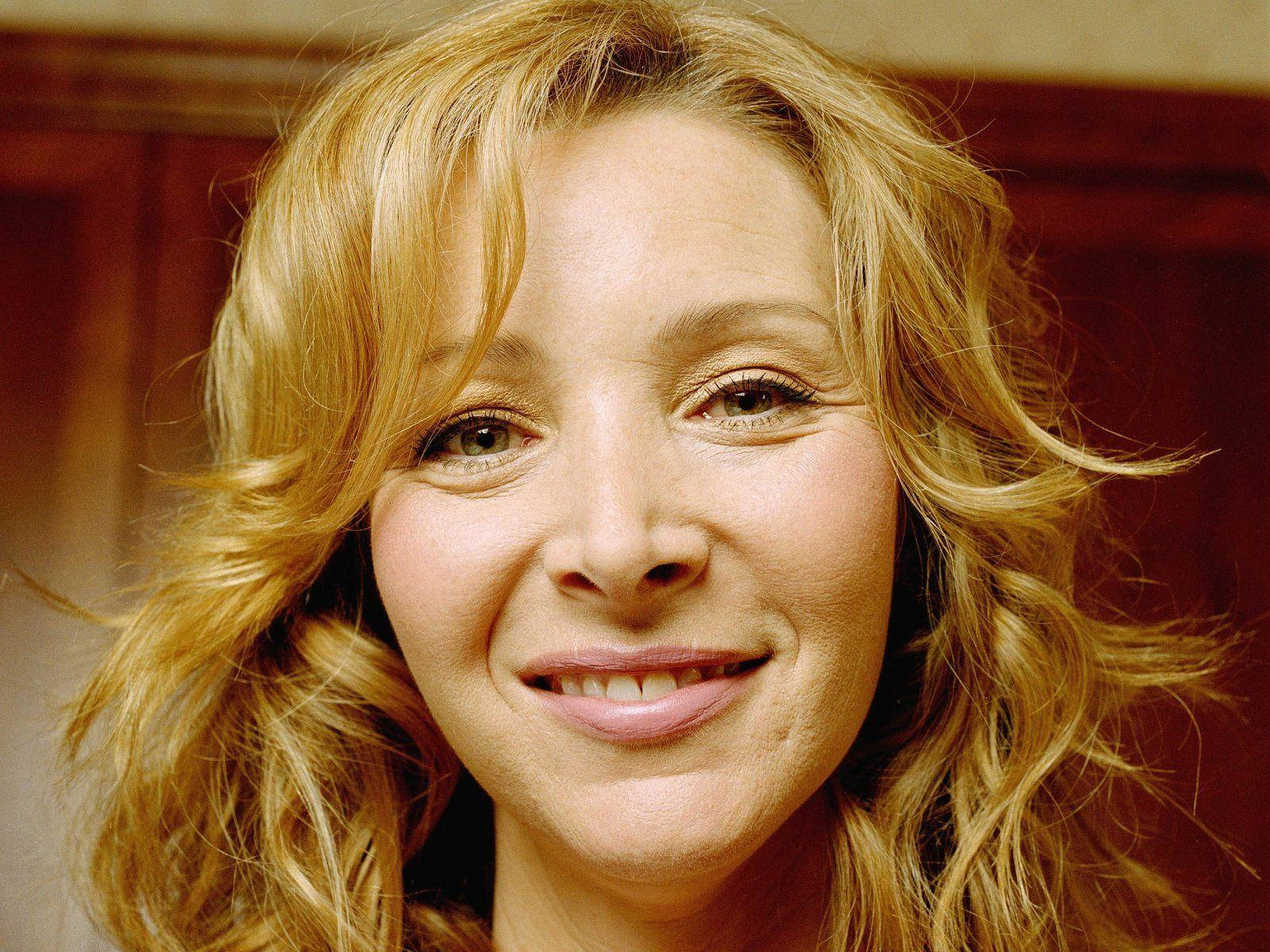 lisa kudrow 桌布 壁紙 — 一個微笑女性的特寫肖像，擁有波浪金髮和溫暖的棕色眼睛，背景為木質。