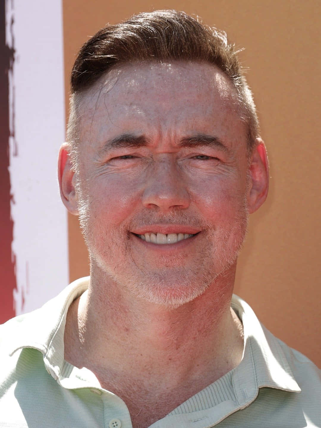 Kevin Durand 桌布 壁紙 — 一個微笑男人的特寫照片，短棕髮和鬍渣，穿著淺綠色有領襯衫，背景是溫暖的橙色和白色。