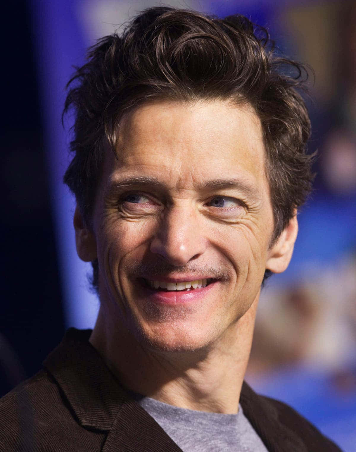 John Hawkes 桌布 壁紙 — 一個微笑男人的特寫肖像，他有深色凌亂的頭髮和藍色眼睛，穿著棕色外套和灰色襯衫。
