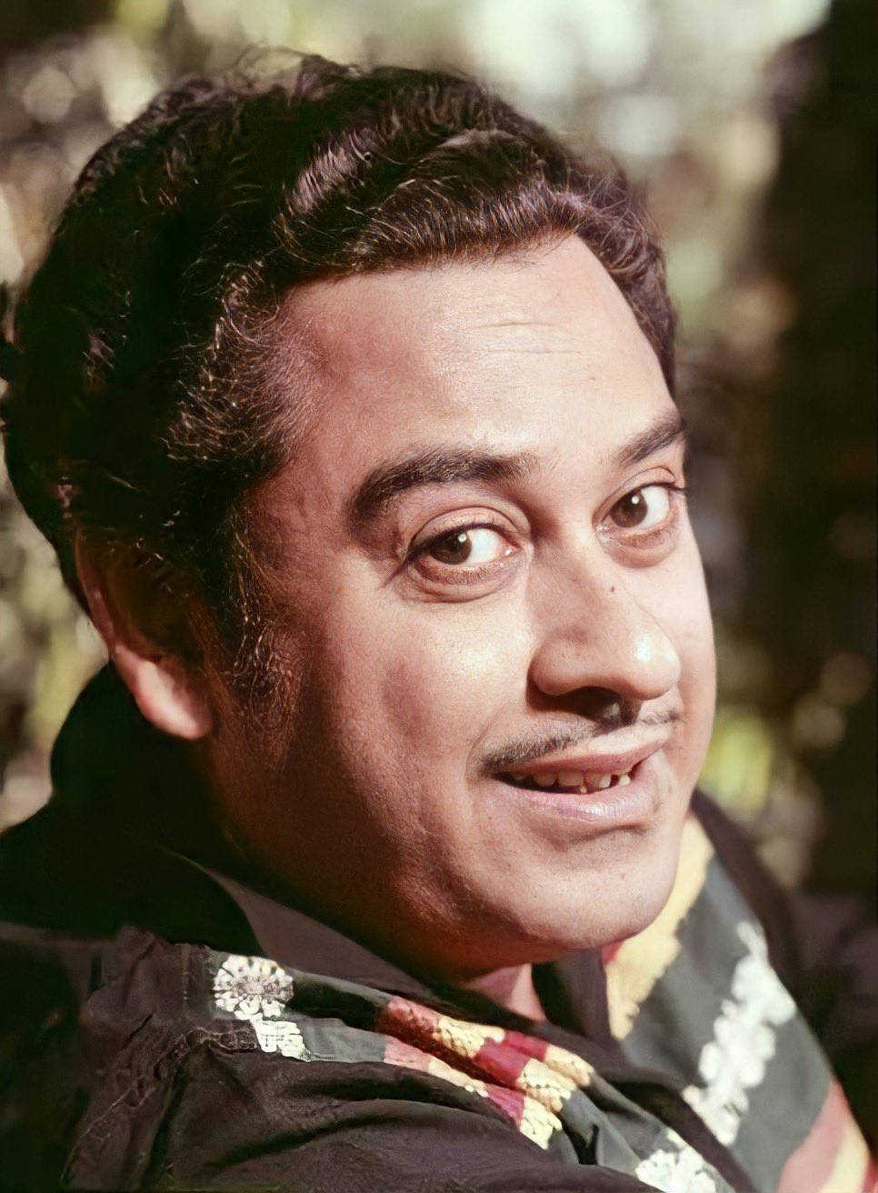 Kishore Kumar 桌布 壁紙 — 一個微笑男人的特寫肖像，他有深色捲髮和鬍子，穿著圖案襯衫，背景是模糊的自然景色。