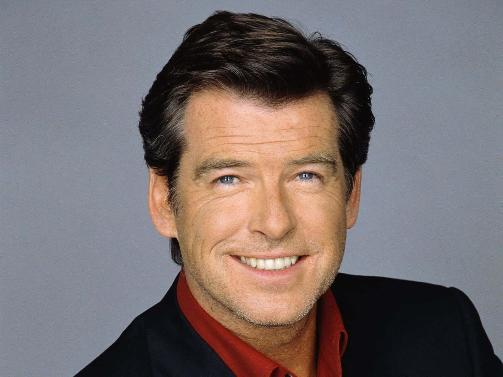pierce brosnan 桌布 壁紙 — 一個微笑男人的特寫肖像，他有藍色眼睛，穿著紅色襯衫和黑色外套，背景是灰色。