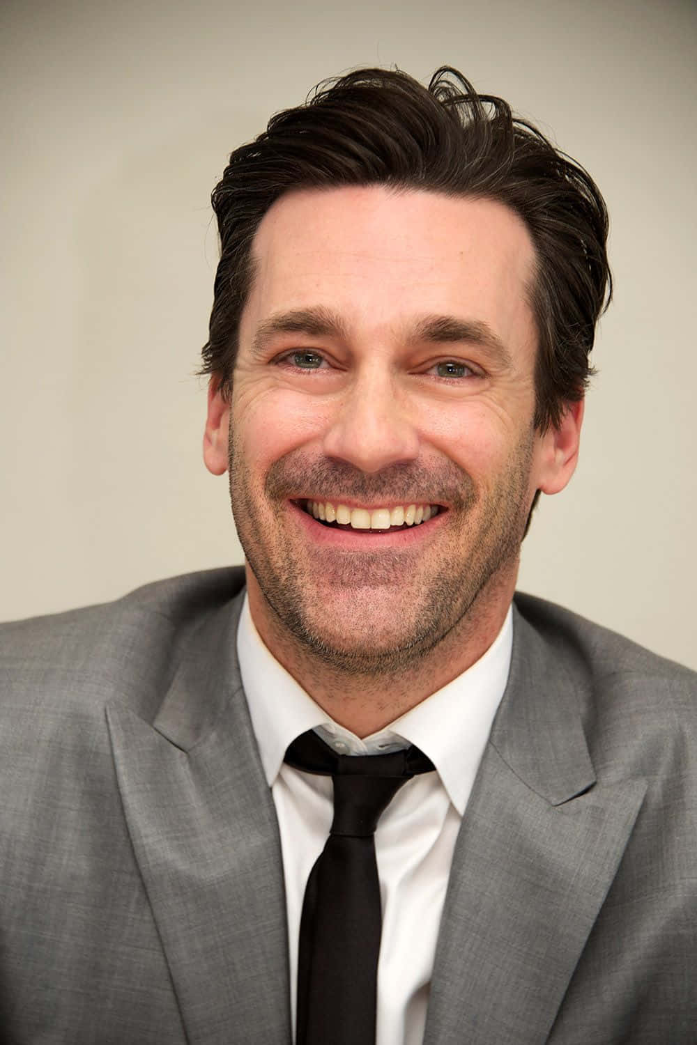 Jon Hamm 桌布 壁紙 — 一個微笑男人的特寫肖像，深色頭髮和鬍渣，穿著灰色西裝和黑色領帶，背景為中性色調。