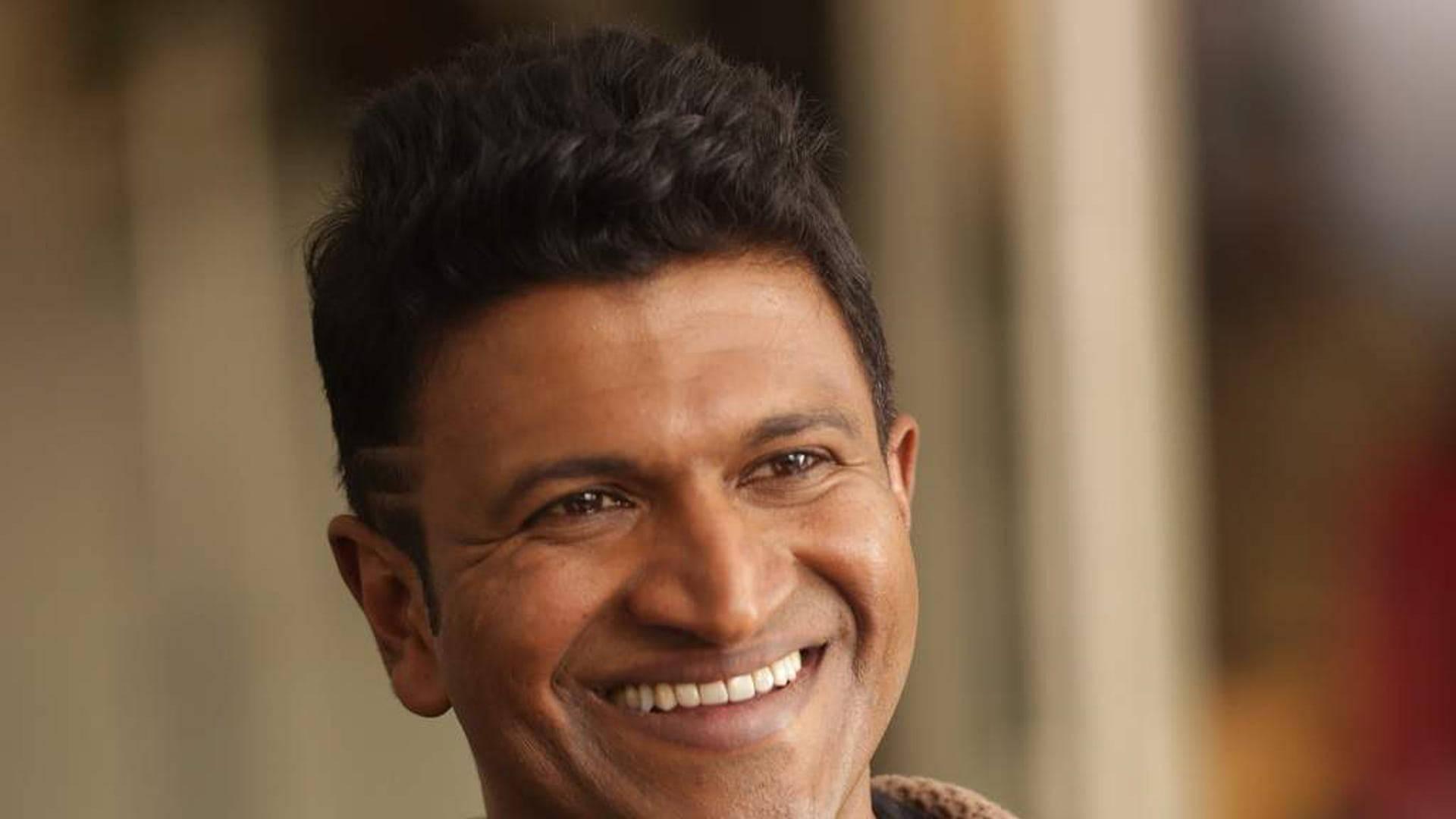 Puneeth Rajkumar 桌布 壁紙 — 一個微笑男士的特寫肖像，深色頭髮，溫暖的眼神，背景柔和模糊。