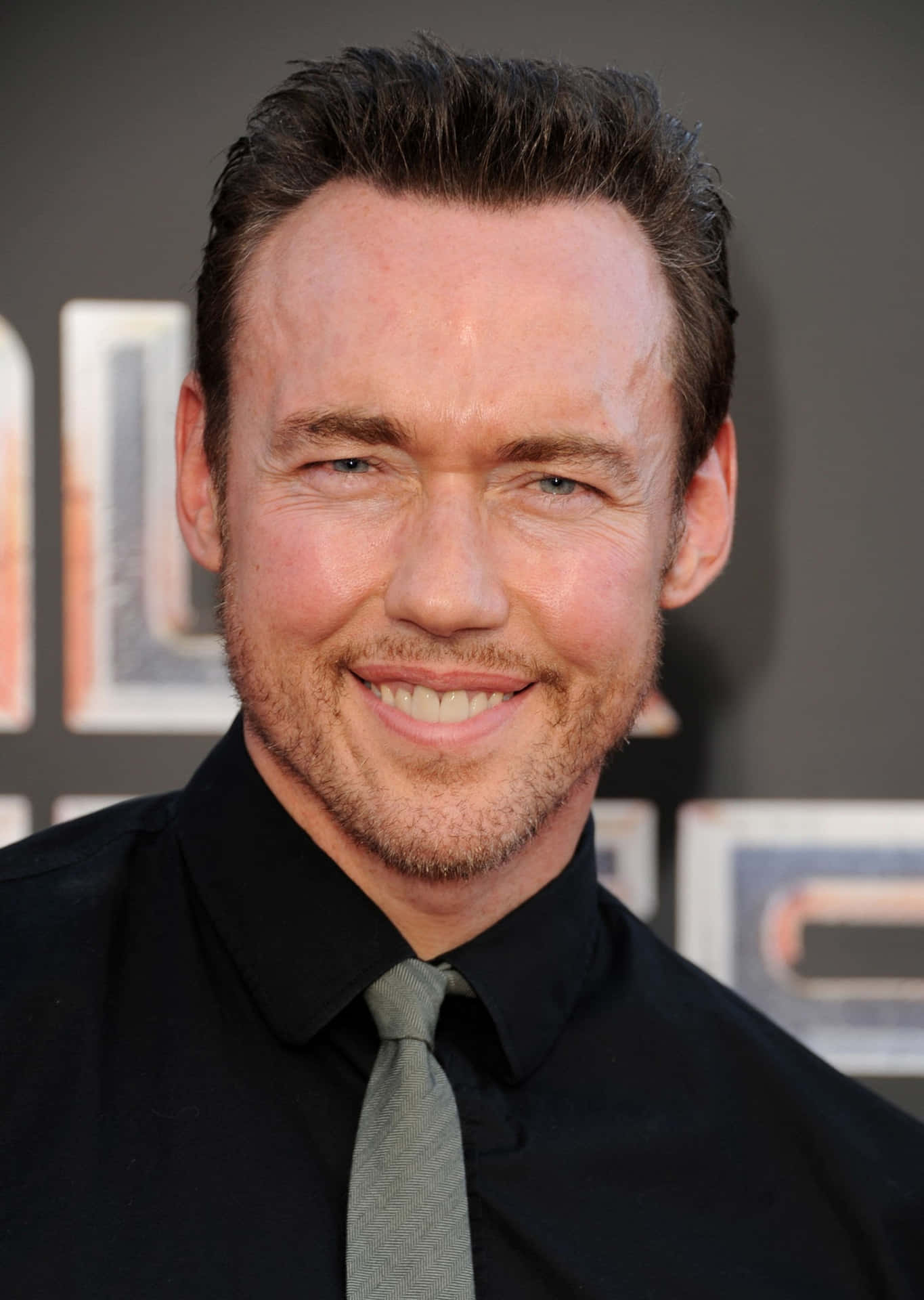 Kevin Durand 桌布 壁紙 — 一個微笑男士的特寫肖像，短黑髮，鬍渣，身穿黑色襯衫和灰色領帶，背景昏暗帶有部分文字。