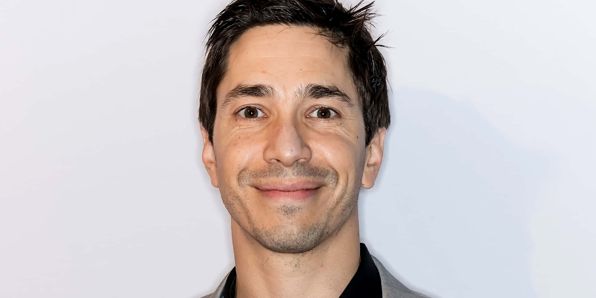 Justin Long 桌布 壁紙 — 一個微笑男子的特寫肖像，深色頭髮和鬍渣，背景為純白色。