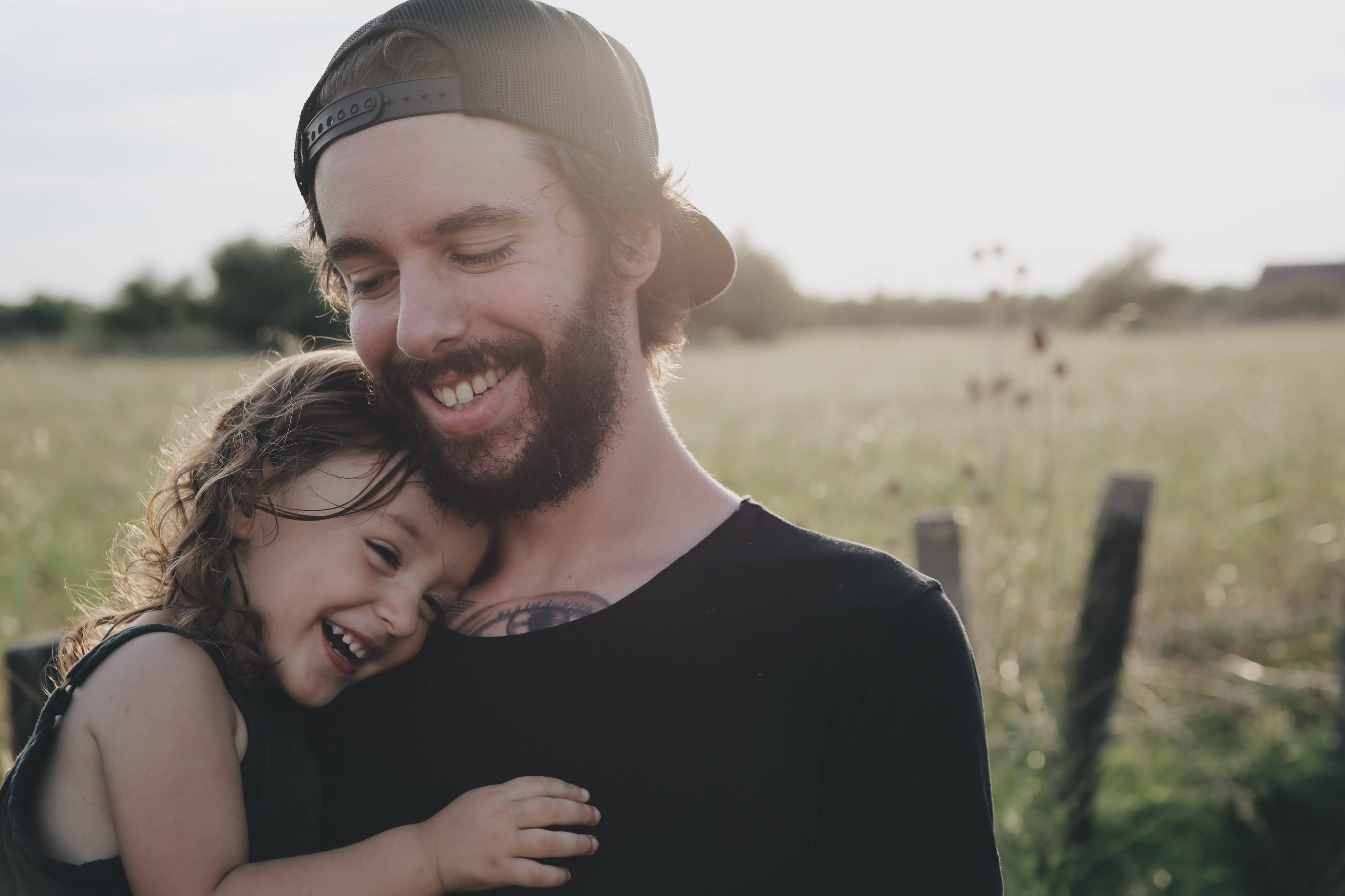 Fathers Day 桌布 壁紙 — 一個微笑留鬍鬚戴帽子的男人在陽光明媚的田野中擁抱著一個大笑的孩子，柔和的金色光線。