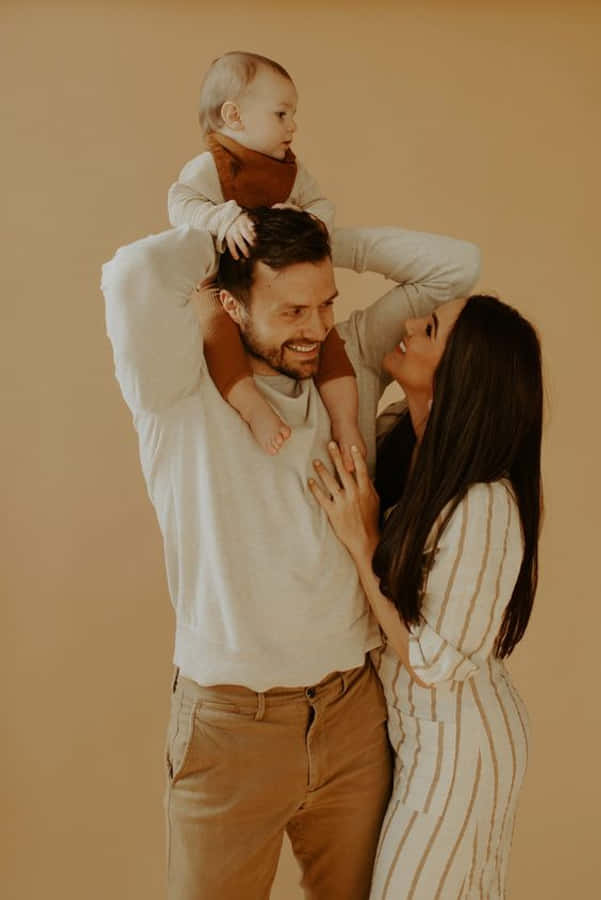 Family Portrait 桌布 壁紙 — 一個微笑的三口之家，寶寶坐在爸爸的肩膀上，在溫暖的米色背景下分享溫馨時刻。