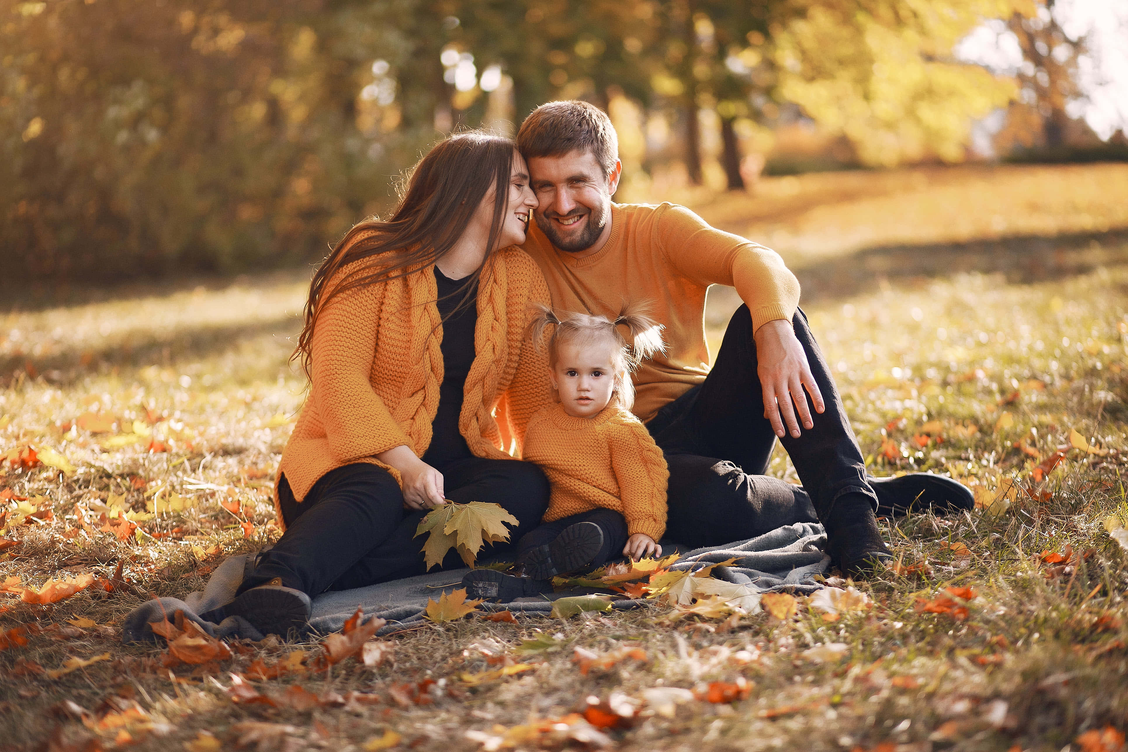 Fall Family 桌布 壁紙 — 一個微笑的三口之家穿著相配的橙色毛衣，坐在陽光普照的公園裡的金色秋葉中的毯子上。