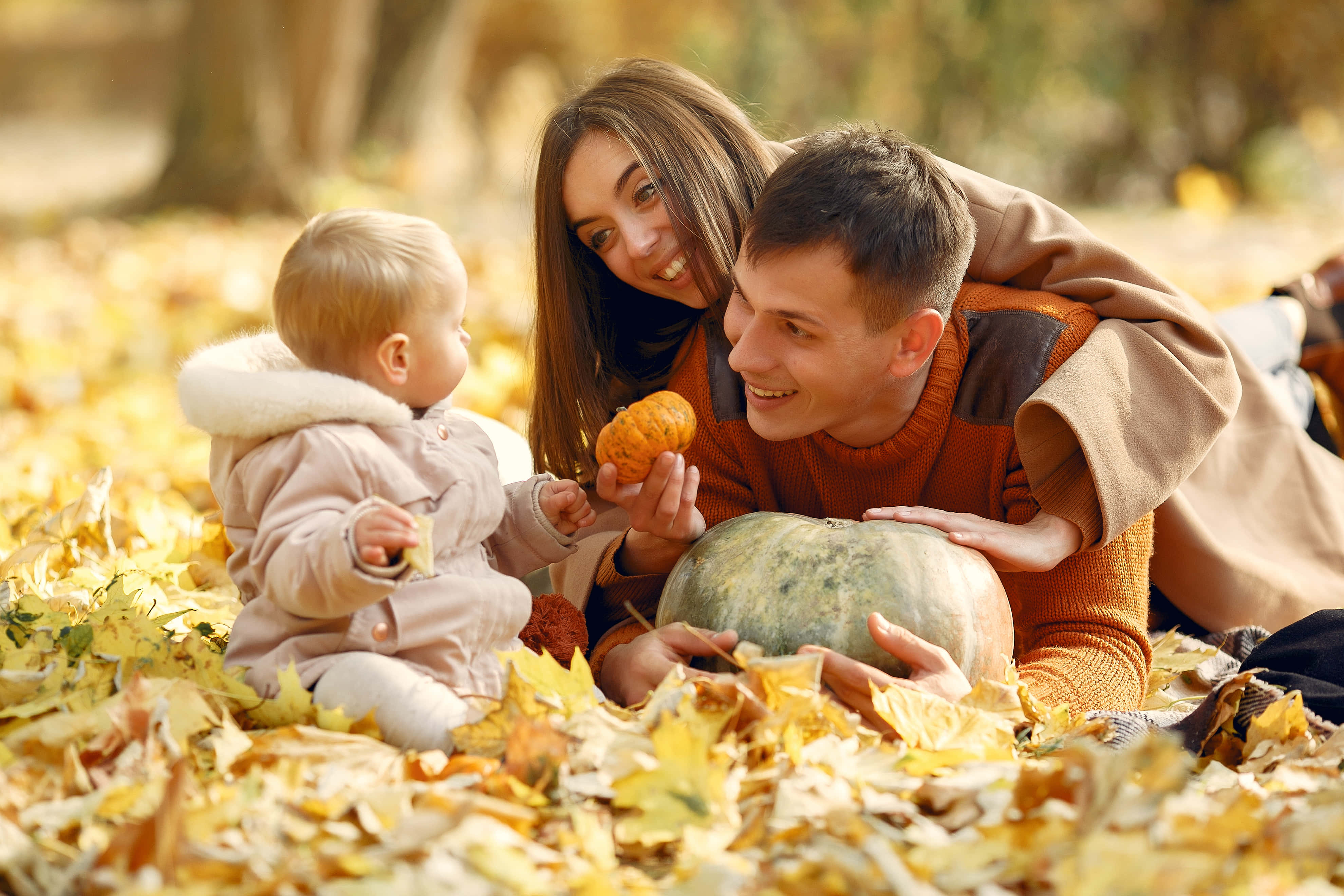Fall Family 桌布 壁紙 — 一個微笑的三口之家躺在陽光普照的公園裡，置身於金黃色的秋葉中，手持南瓜。