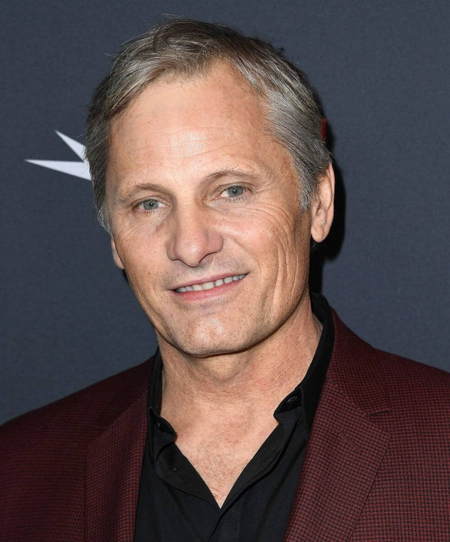 actor 桌布 壁紙 — 一個微笑的中年男子特寫肖像，灰白頭髮，身穿棗紅色西裝外套配黑色襯衫，背景為深藍色並帶有白色星星標誌。