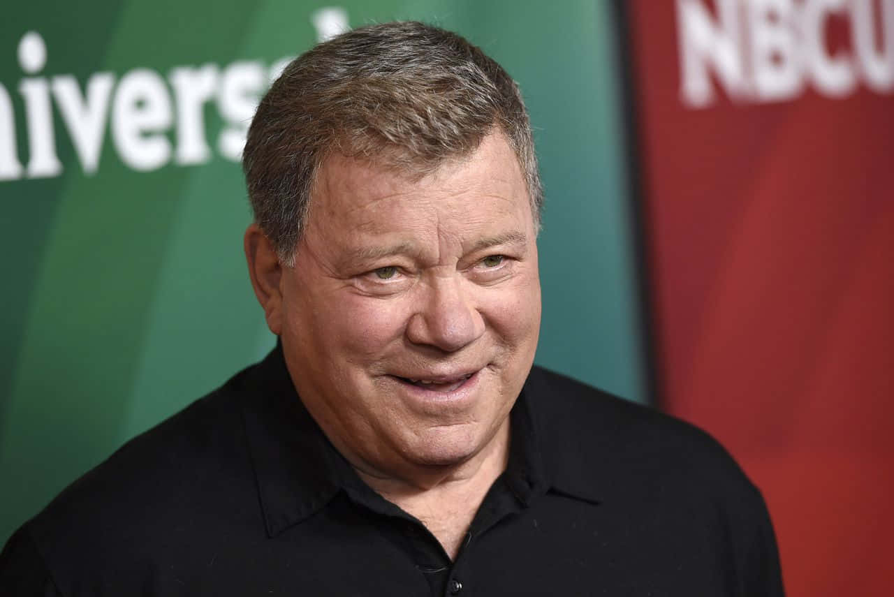 william shatner 桌布 壁紙 — 一個微笑的中年男子身穿黑色襯衫，以綠色和紅色品牌背景的特寫肖像。