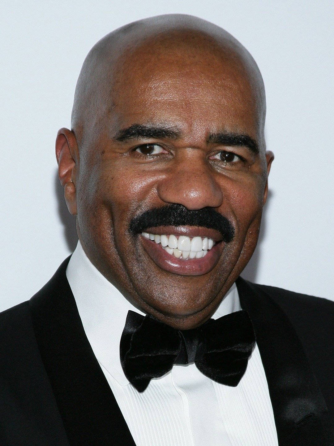 steve harvey 桌布 壁紙 — 一個微笑的光頭男人特寫肖像，留著濃密的鬍子，穿著黑色燕尾服和領結，背景是純白色。