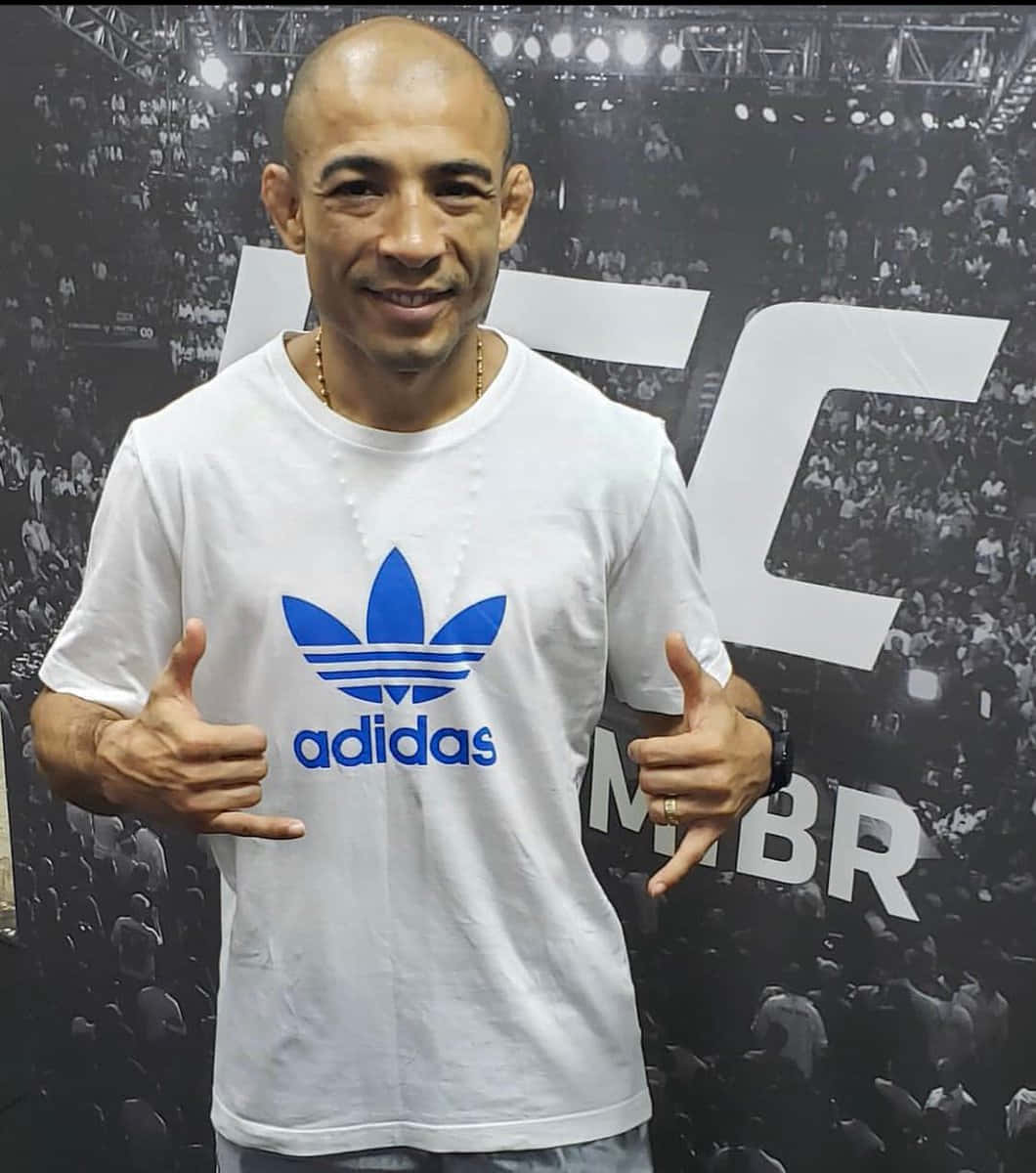 UFC 桌布 壁紙 — 一個微笑的光頭男人穿著白色Adidas T恤，在UFC活動背景前豎起兩個大拇指，背景有群眾。