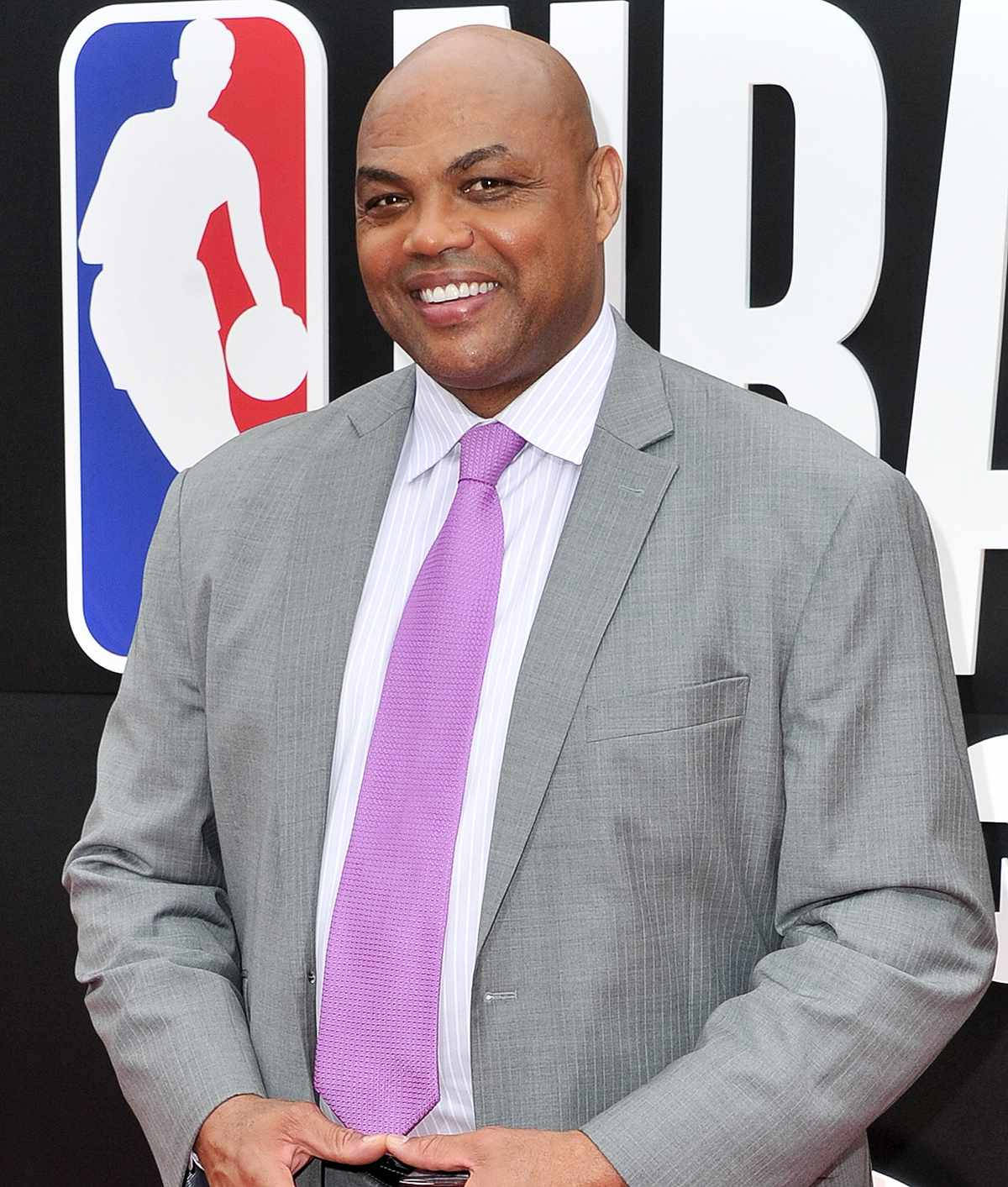 Charles Barkley 桌布 壁紙 — 一個微笑的光頭男士，身穿灰色西裝和紫色領帶，自信地站在大型NBA標誌背景前。