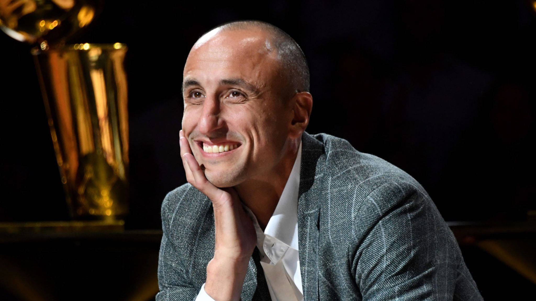 Manu 桌布 壁紙 — 一個微笑的光頭男士穿著灰色西裝，在深色背景前將下巴擱在手上，旁邊有一個金色獎盃。