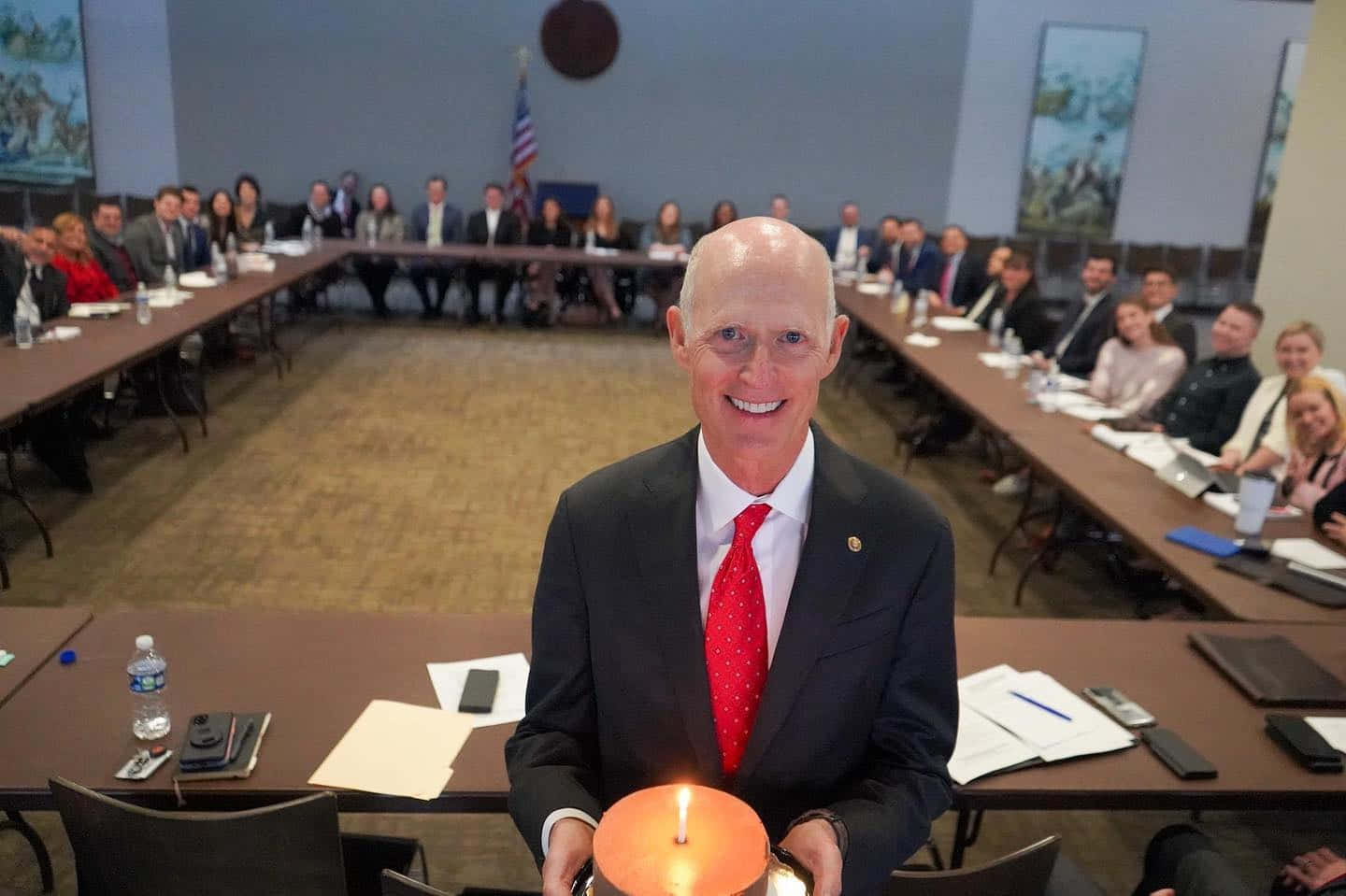 Birthday 桌布 壁紙 — 一個微笑的光頭男士穿著西裝，在會議桌上拿著一支點燃的蠟燭，背景有觀眾坐著。