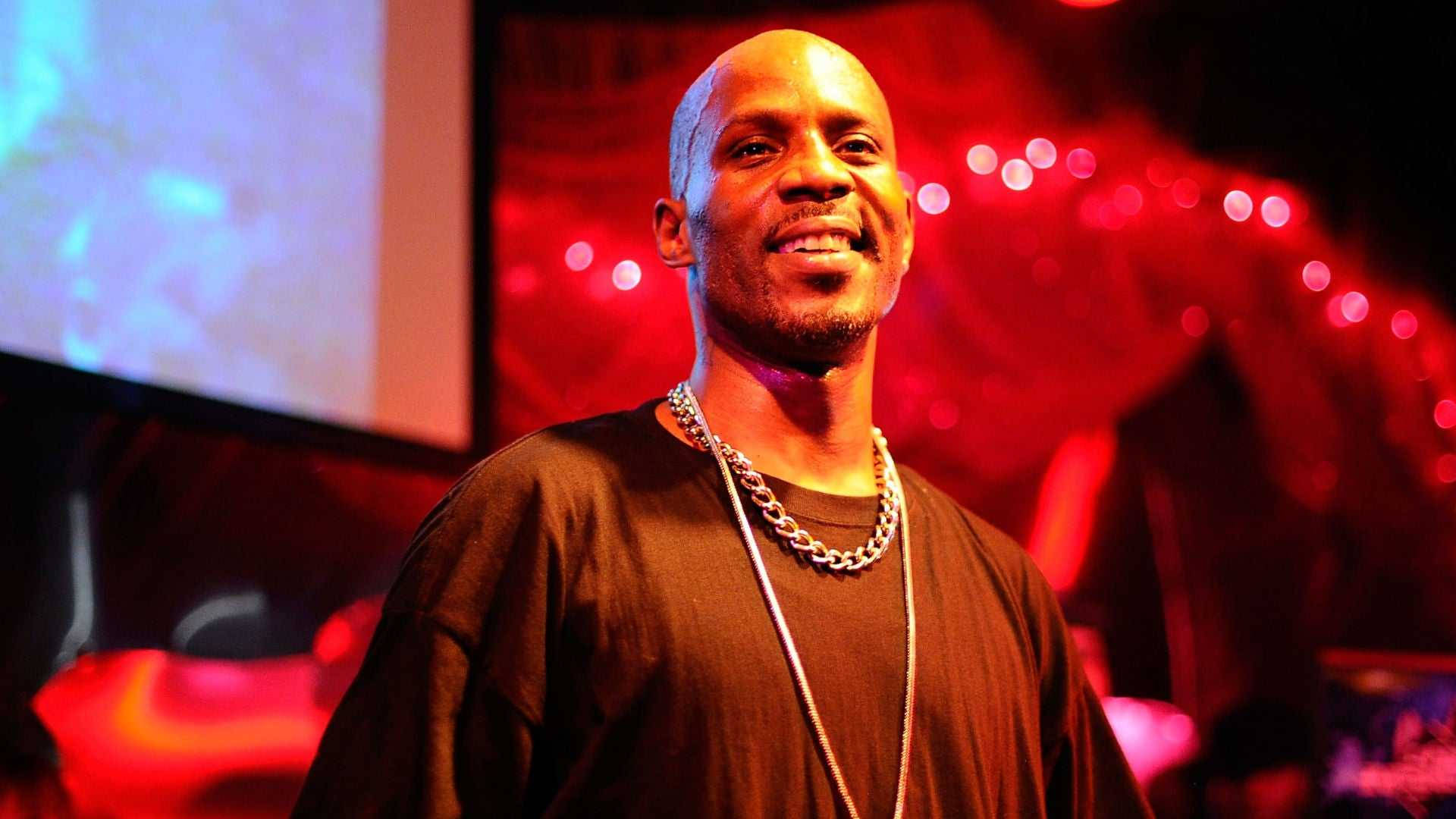 Dmx 桌布 壁紙 — 一個微笑的光頭男子，身穿黑色襯衫和銀色鏈條，站在被紅色和藍色燈光籠罩的舞台上。