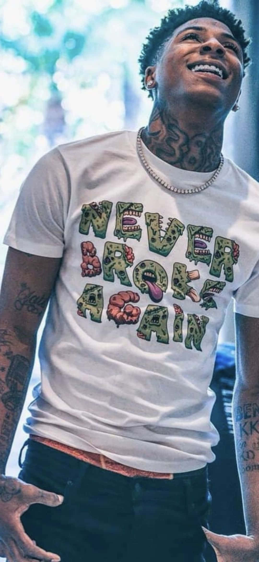 Youngboy 桌布 壁紙 — 一個微笑的刺青男子，身穿印有殭屍主題文字的白色圖案T恤，對著明亮模糊的背景向上大笑。