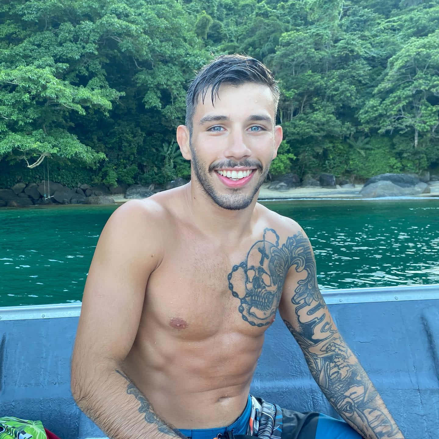 Lake 桌布 壁紙 — 一個微笑的刺青男子赤裸上身坐在船上，背景是碧綠的海水和茂密的綠色叢林。