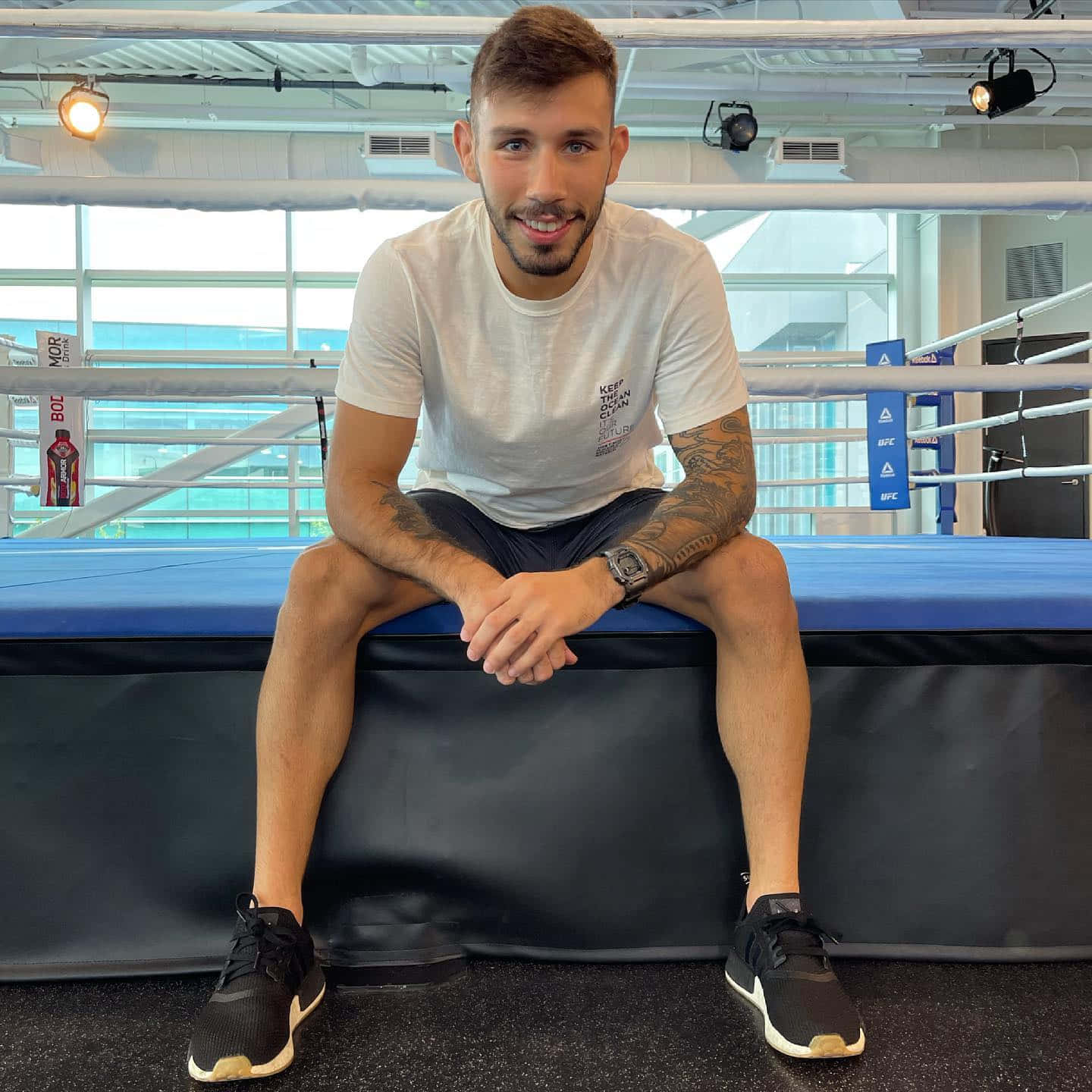 MMA 桌布 壁紙 — 一個微笑的刺青男子隨意地坐在明亮健身房的拳擊台角墊上。