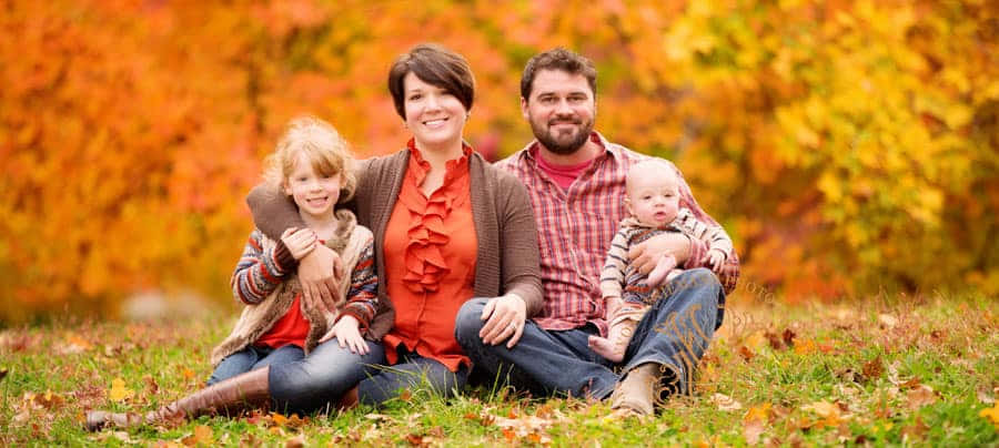 Fall Family 桌布 壁紙 — 一個微笑的四口之家坐在草地上，周圍環繞著充滿活力的橙色秋葉。