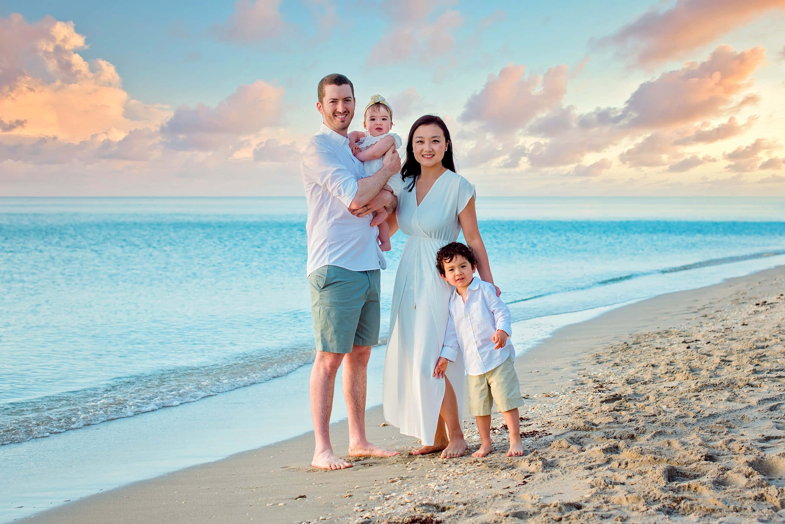 Beach Family 桌布 壁紙 — 一個微笑的四口之家赤腳站在沙灘上，背景是日落時分的柔和波浪和粉彩雲彩。