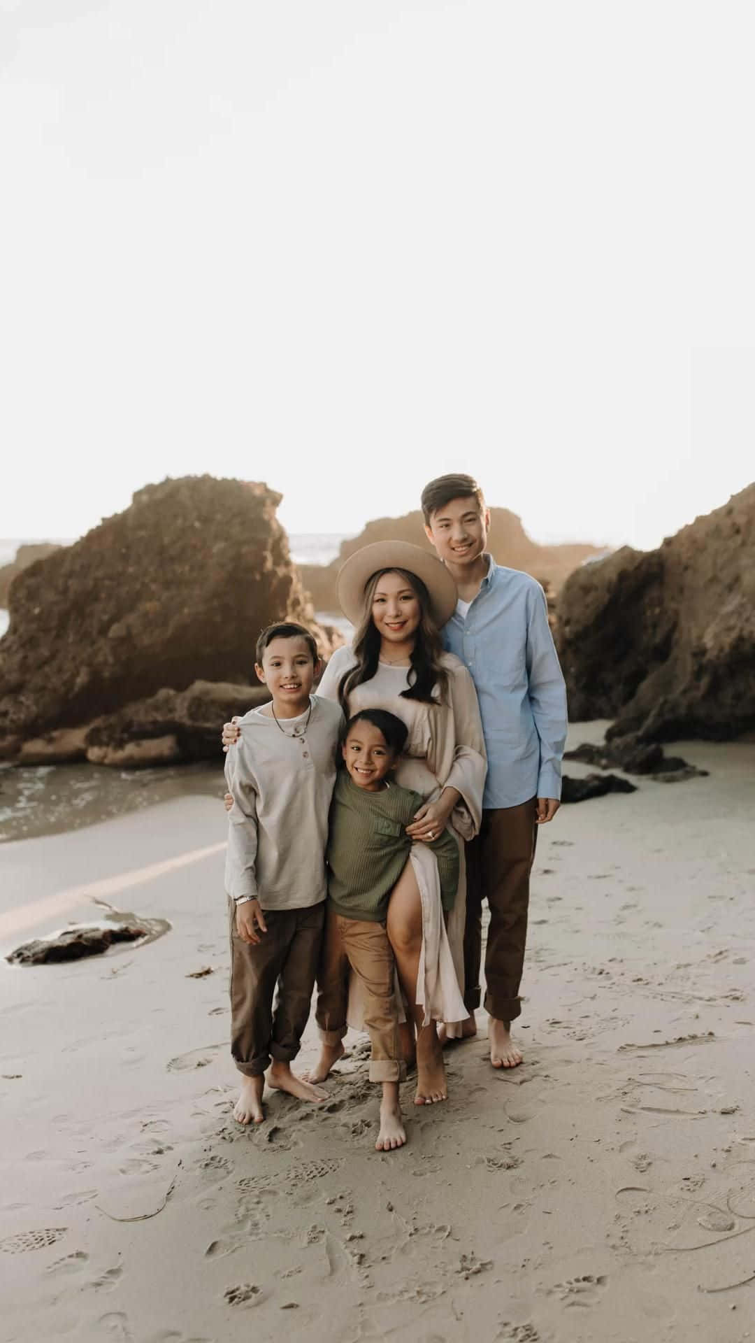 Family Beach 桌布 壁紙 — 一個微笑的四口之家赤腳站在沙灘上，身後有巨大的岩石，沐浴在柔和的金色光線中。