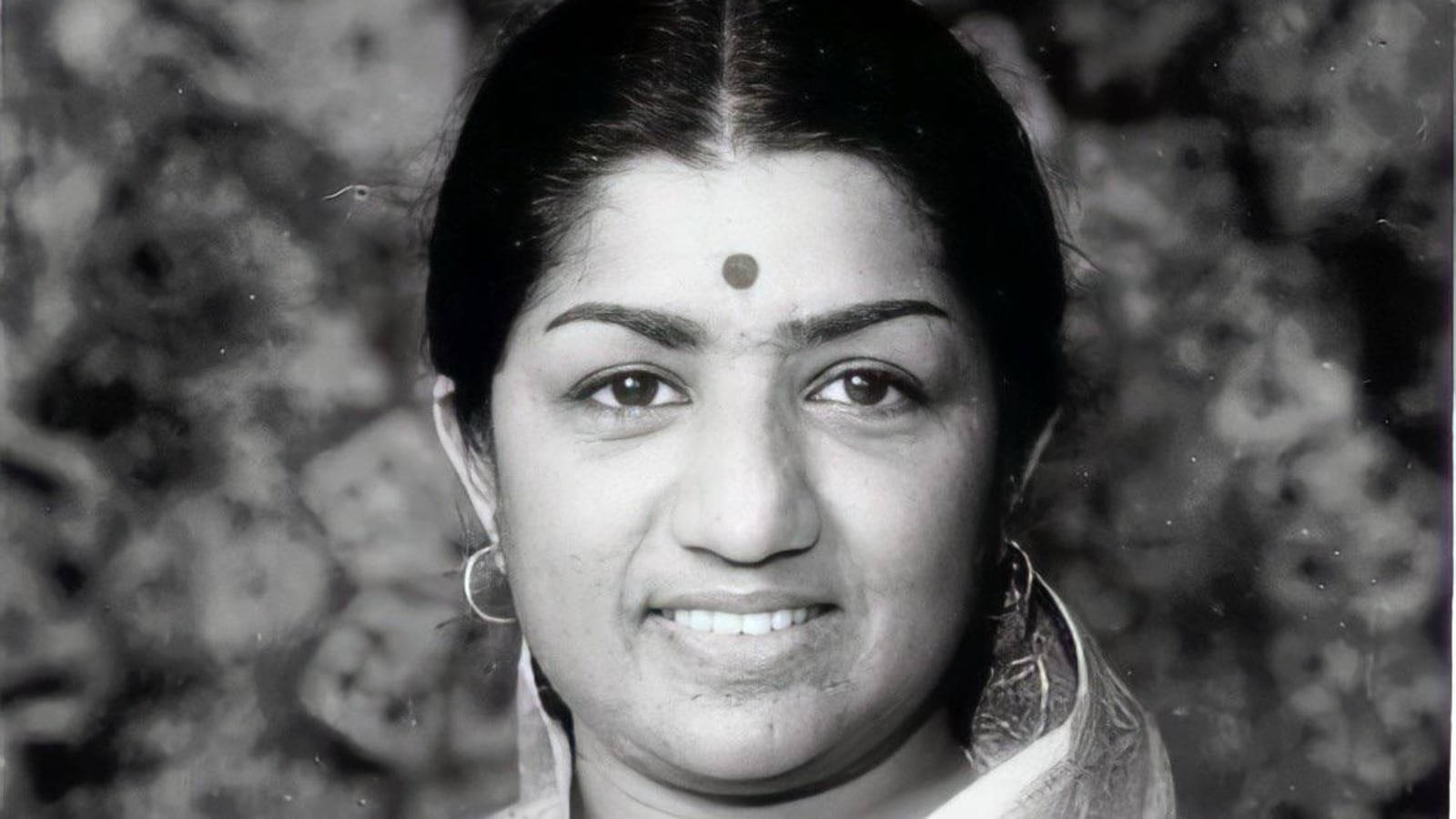 Lata Mangeshkar 桌布 壁紙 — 一個微笑的女人，額頭上有吉祥痣，戴著圓環耳環，在紋理背景前的黑白肖像。