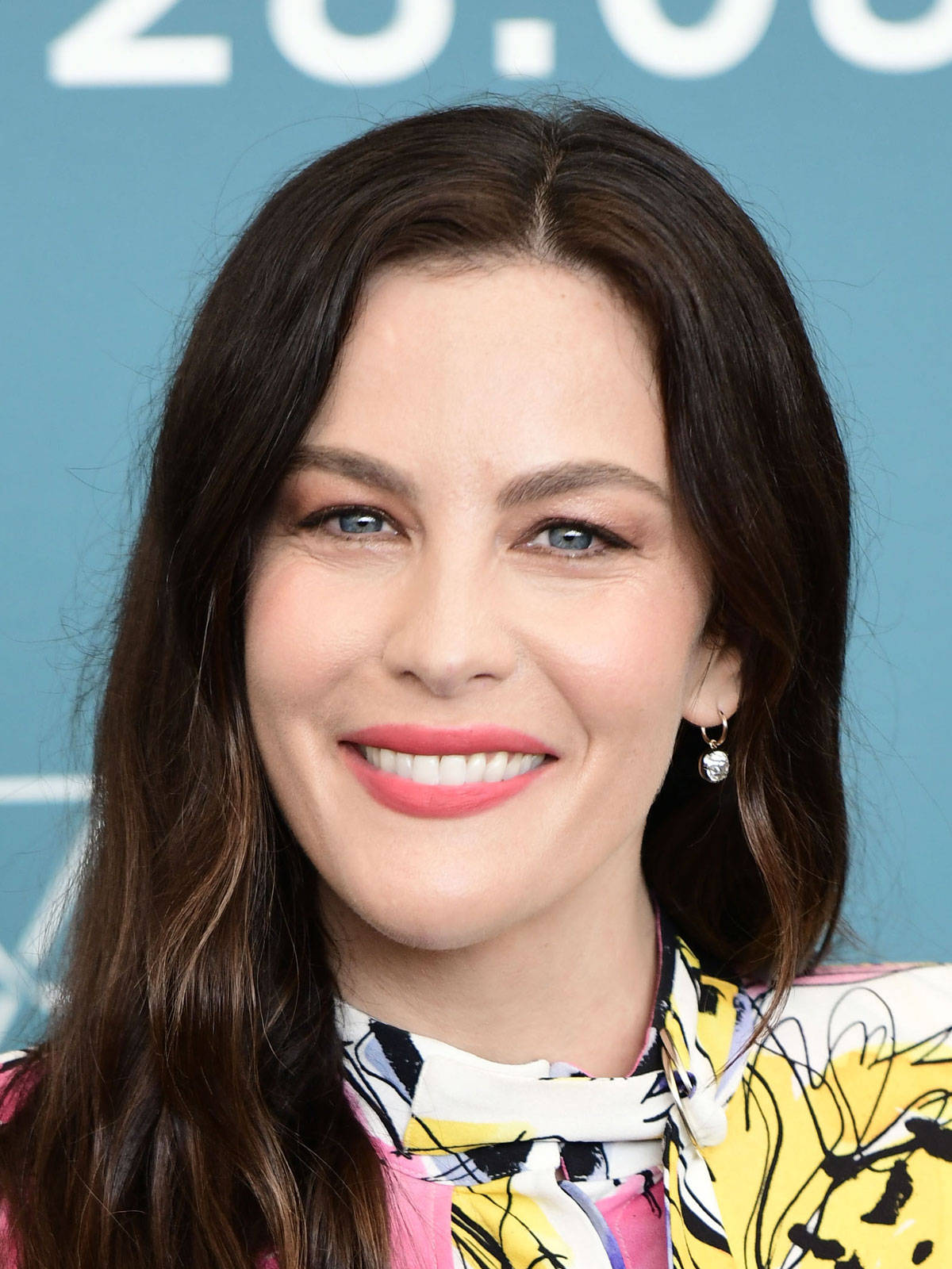 Liv Tyler 桌布 壁紙 — 一個微笑的女人特寫肖像，她擁有深色頭髮和藍色眼睛，戴著一條色彩繽紛的花卉圍巾，背景是藍色。