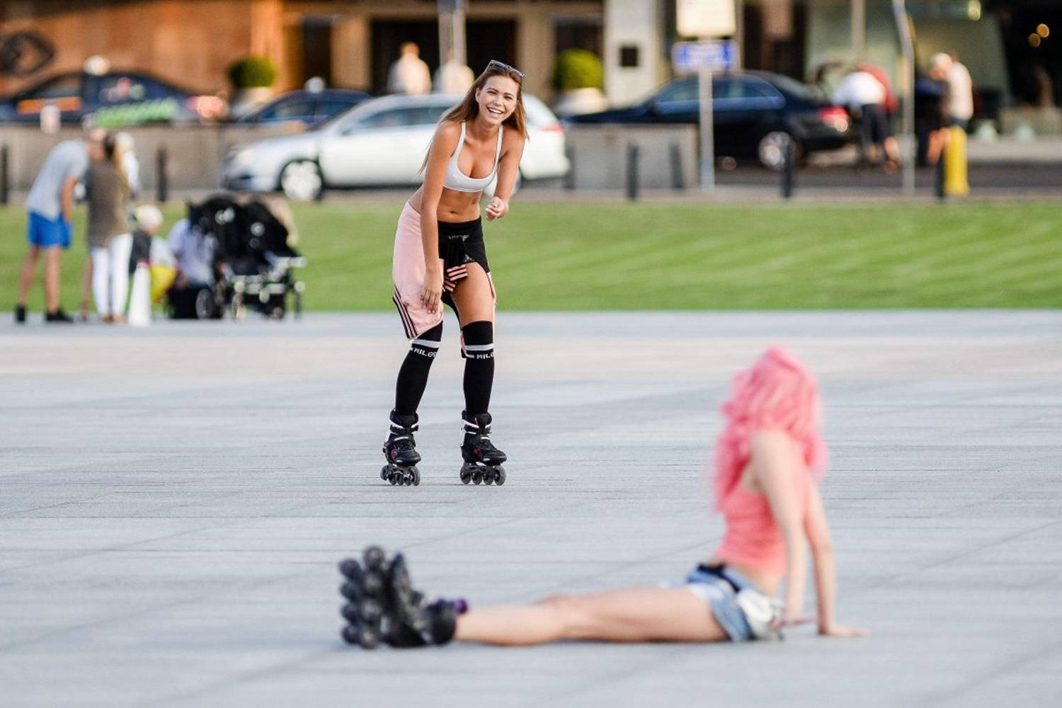 skating 桌布 壁紙 — 一個微笑的女人穿著溜冰鞋在鋪砌的廣場上滑行，而另一個粉紅色頭髮的人在附近伸展。