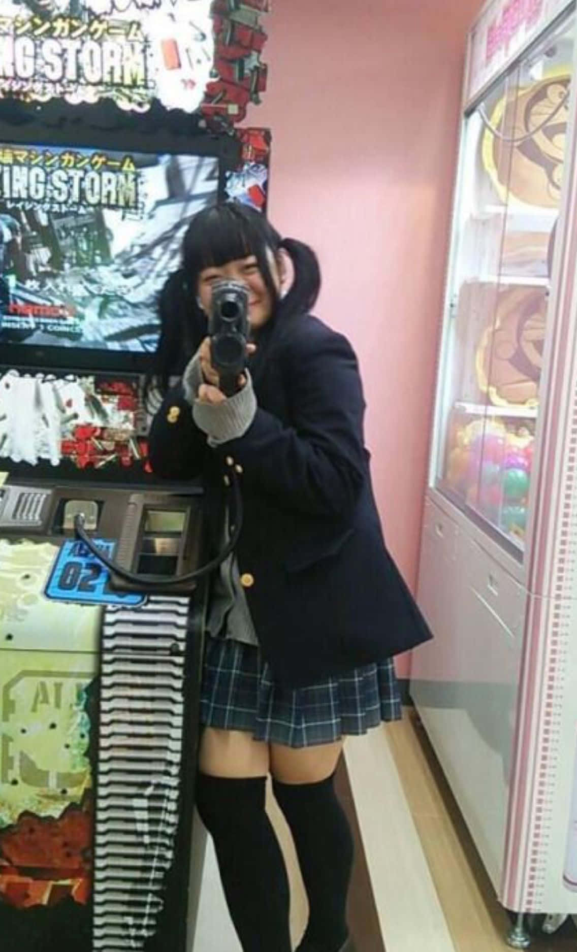 垃圾美學 桌布 壁紙 — 一個微笑的女孩穿著校服，在射擊街機遊戲中頑皮地擺弄著槍形控制器。