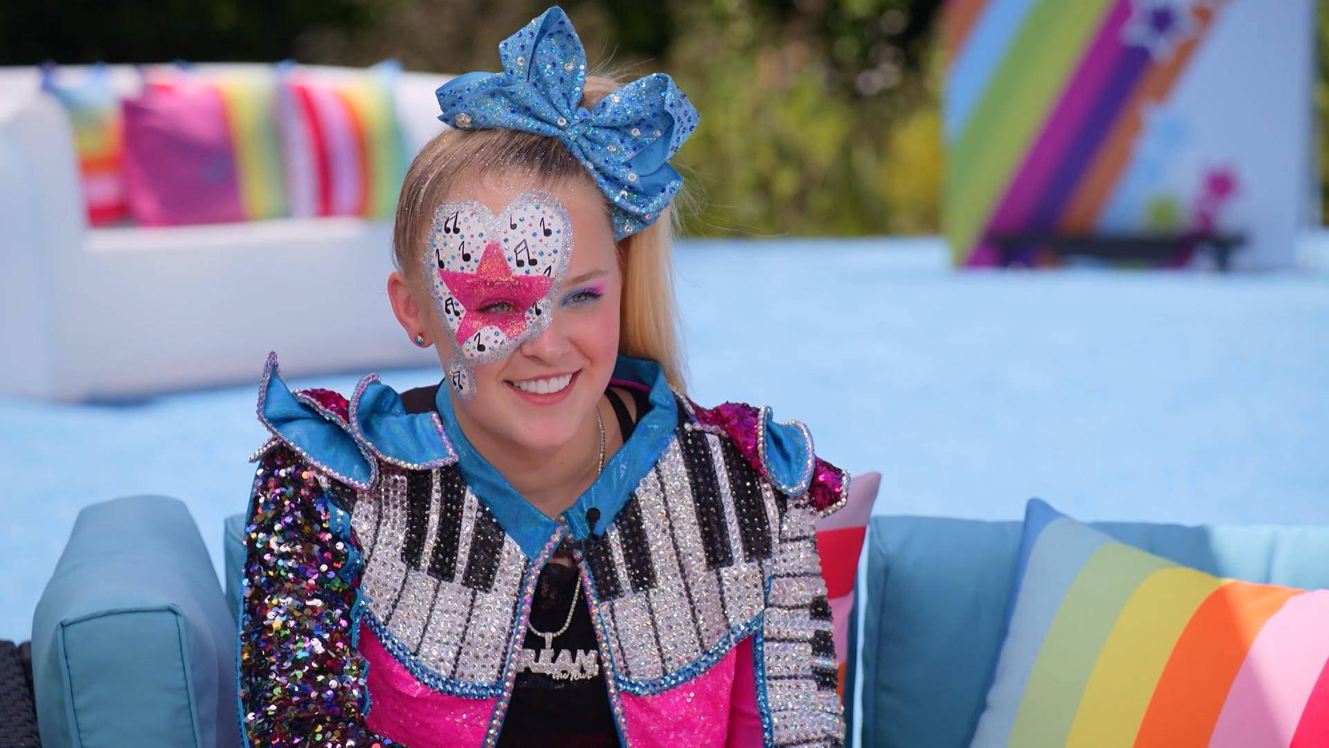 JoJo Siwa 桌布 壁紙 — 一個微笑的女孩穿著閃閃發光的鋼琴鍵外套和藍色蝴蝶結，坐在帶有彩虹枕頭的彩色沙發上。