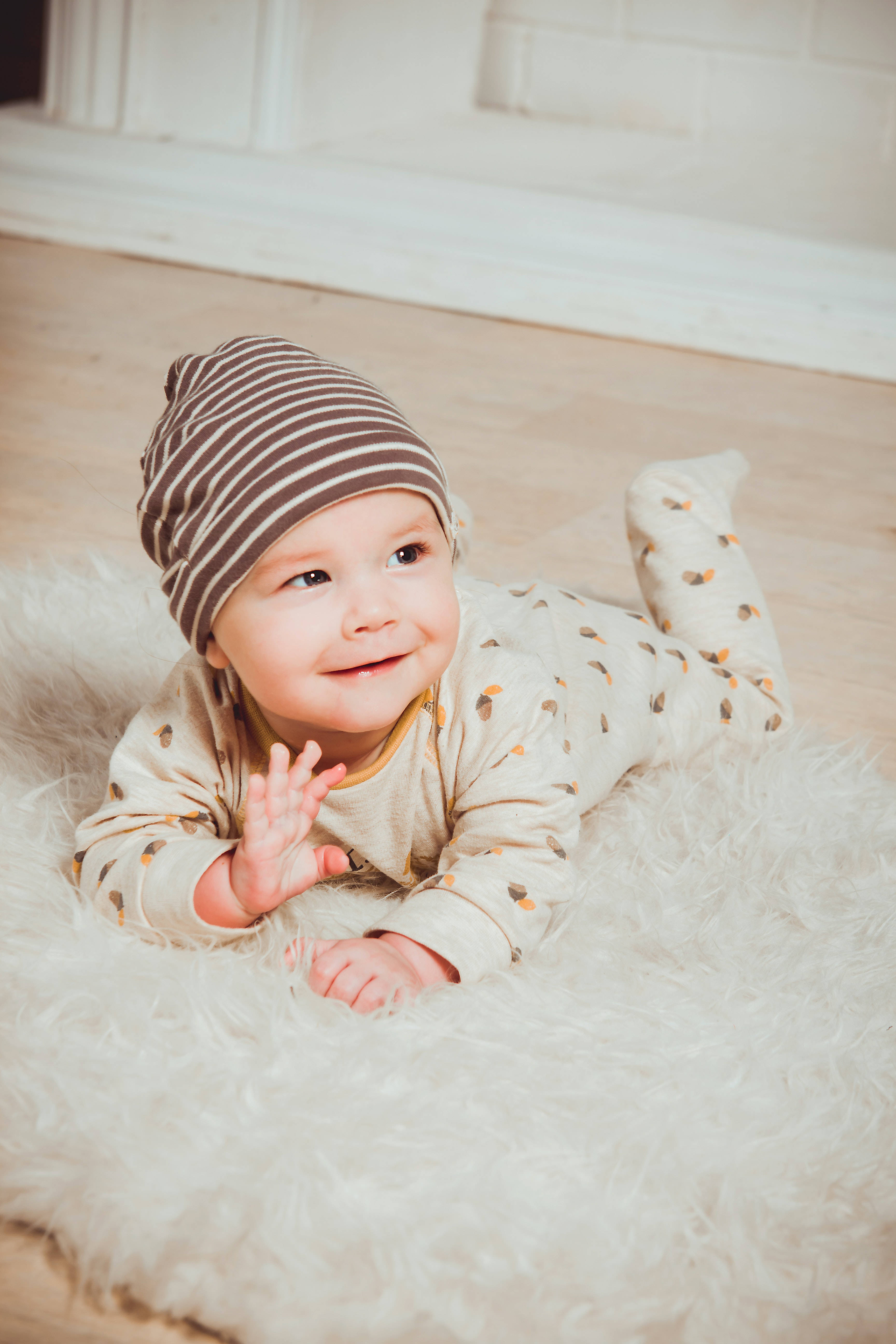 Baby Boy 桌布 壁紙 — 一個微笑的嬰兒戴著條紋帽子，躺在室內柔軟的白色地毯上，開心地向鏡頭揮手。