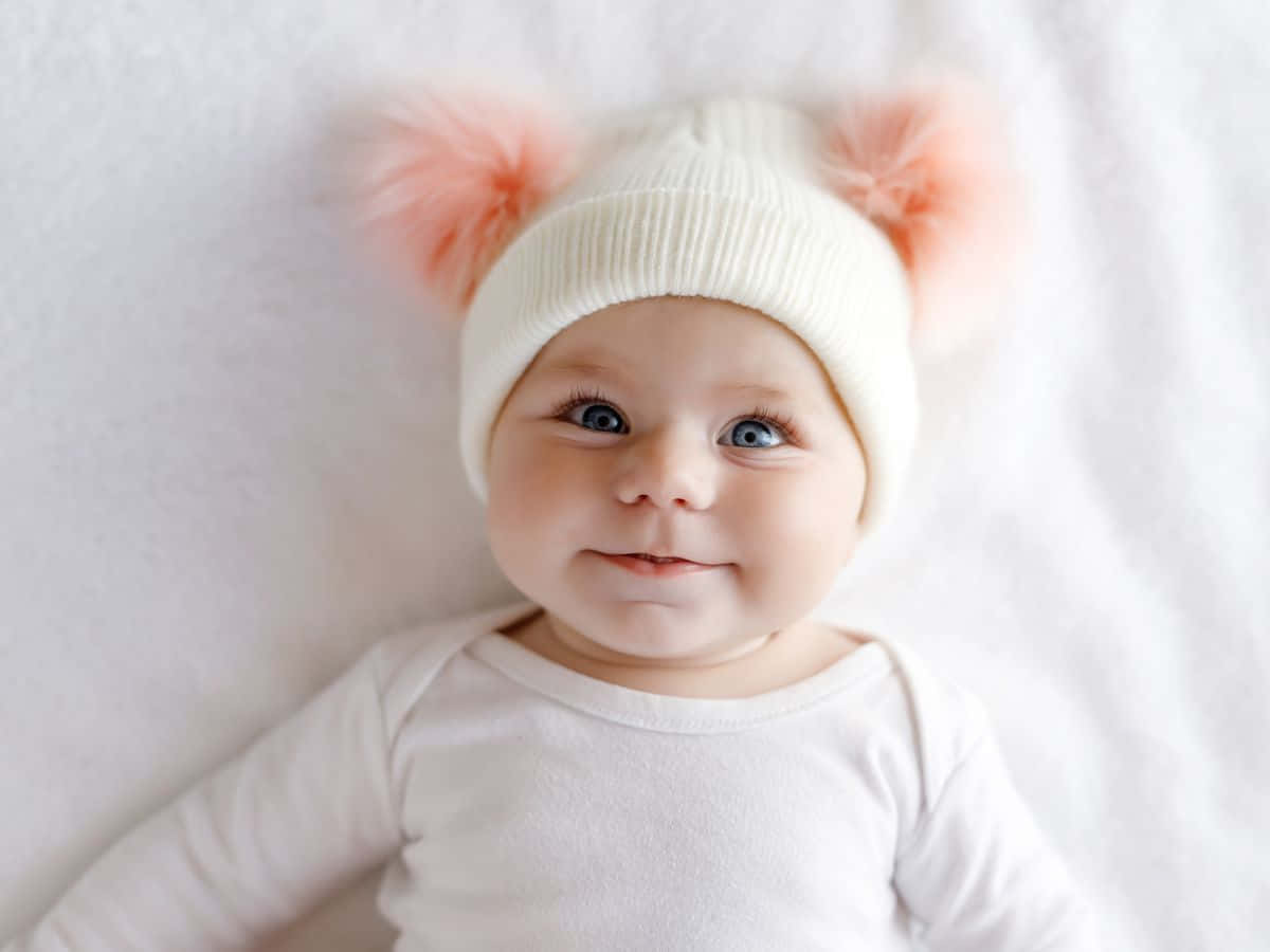 Baby Girl 桌布 壁紙 — 一個微笑的嬰兒戴著白色針織帽，帽上有粉紅色絨球，躺在柔軟的白色毯子上。