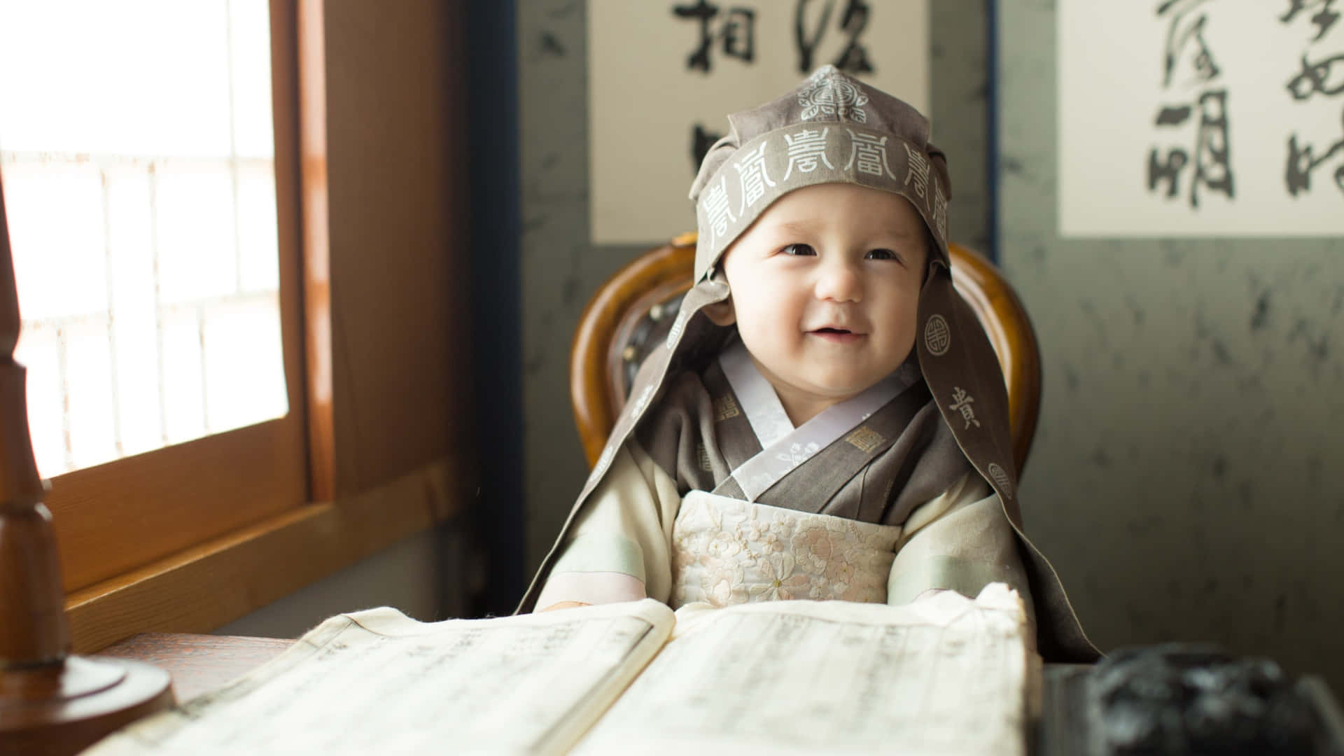 smiling 桌布 壁紙 — 一個微笑的嬰兒穿著傳統韓國韓服，坐在陽光充足的書房的木桌上，桌上放著一本打開的書和硯台。