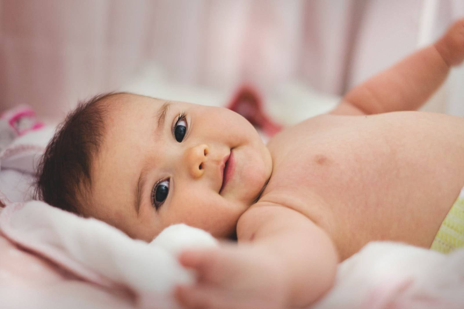 Baby Boy 桌布 壁紙 — 一個微笑的嬰兒近距離特寫，躺在柔軟的粉紅色毯子上，有著好奇的深色眼睛和胖嘟嘟的臉頰。